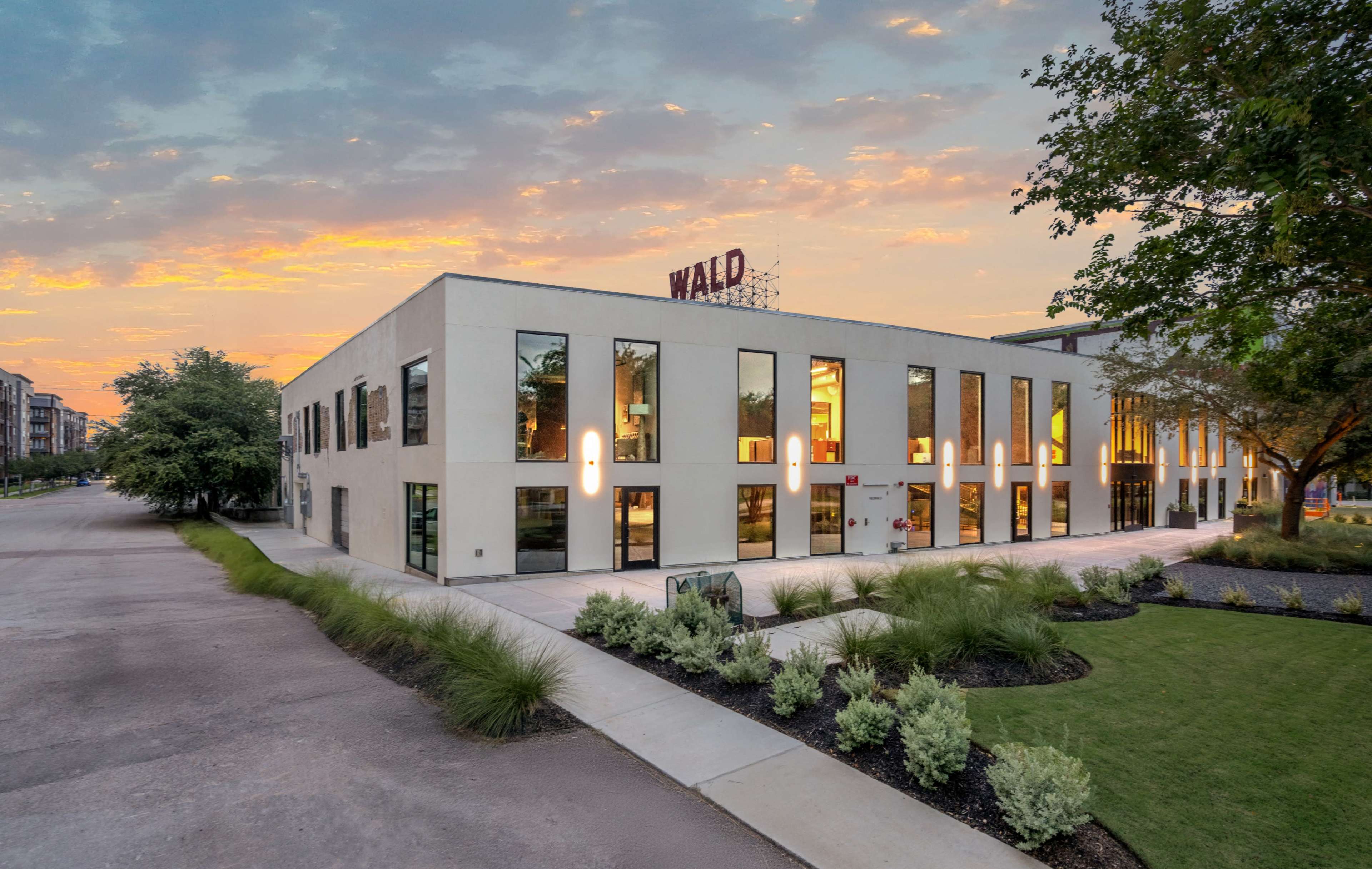 The image shows a modern building with large windows and a sign reading "Wald" on the roof, surrounded by landscaped greenery at sunset.