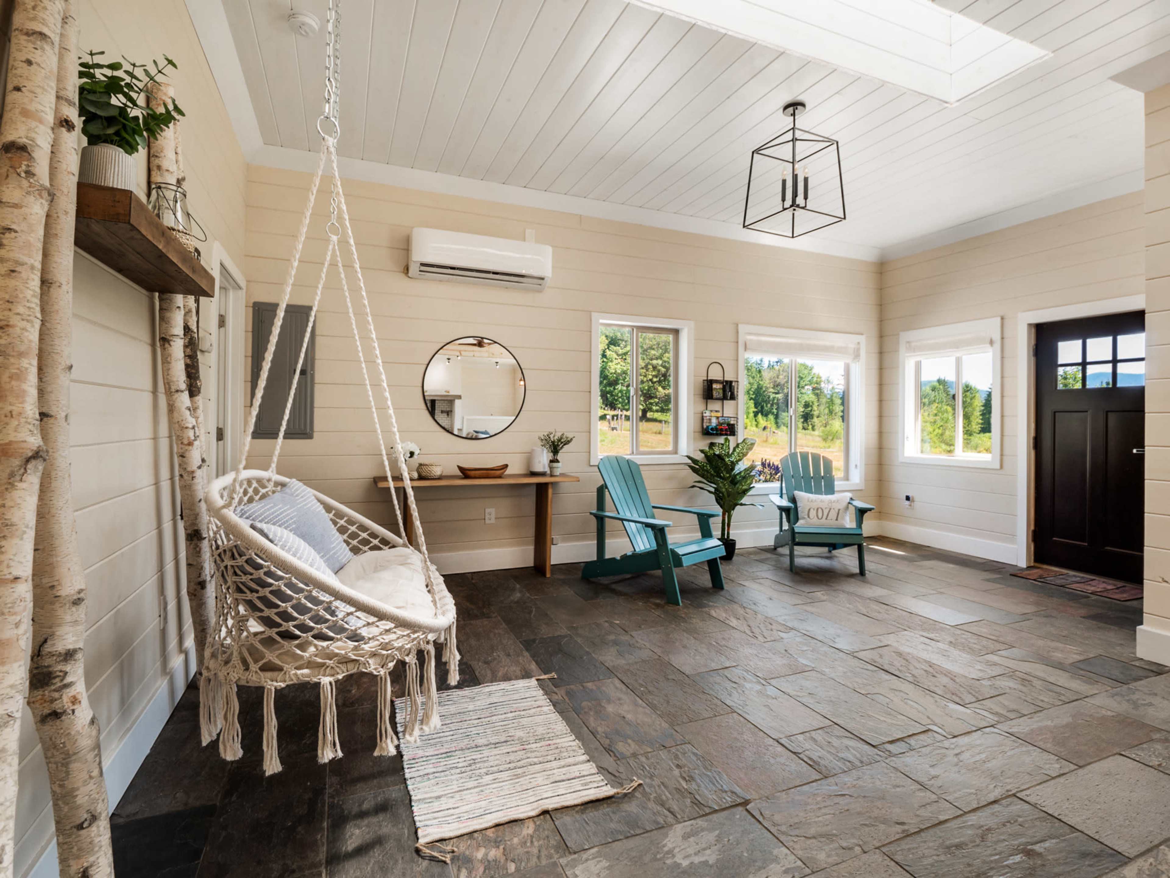 A bright interior features a hanging swing chair, two blue Adirondack chairs, and a slate floor, illuminated by natural light from large windows.