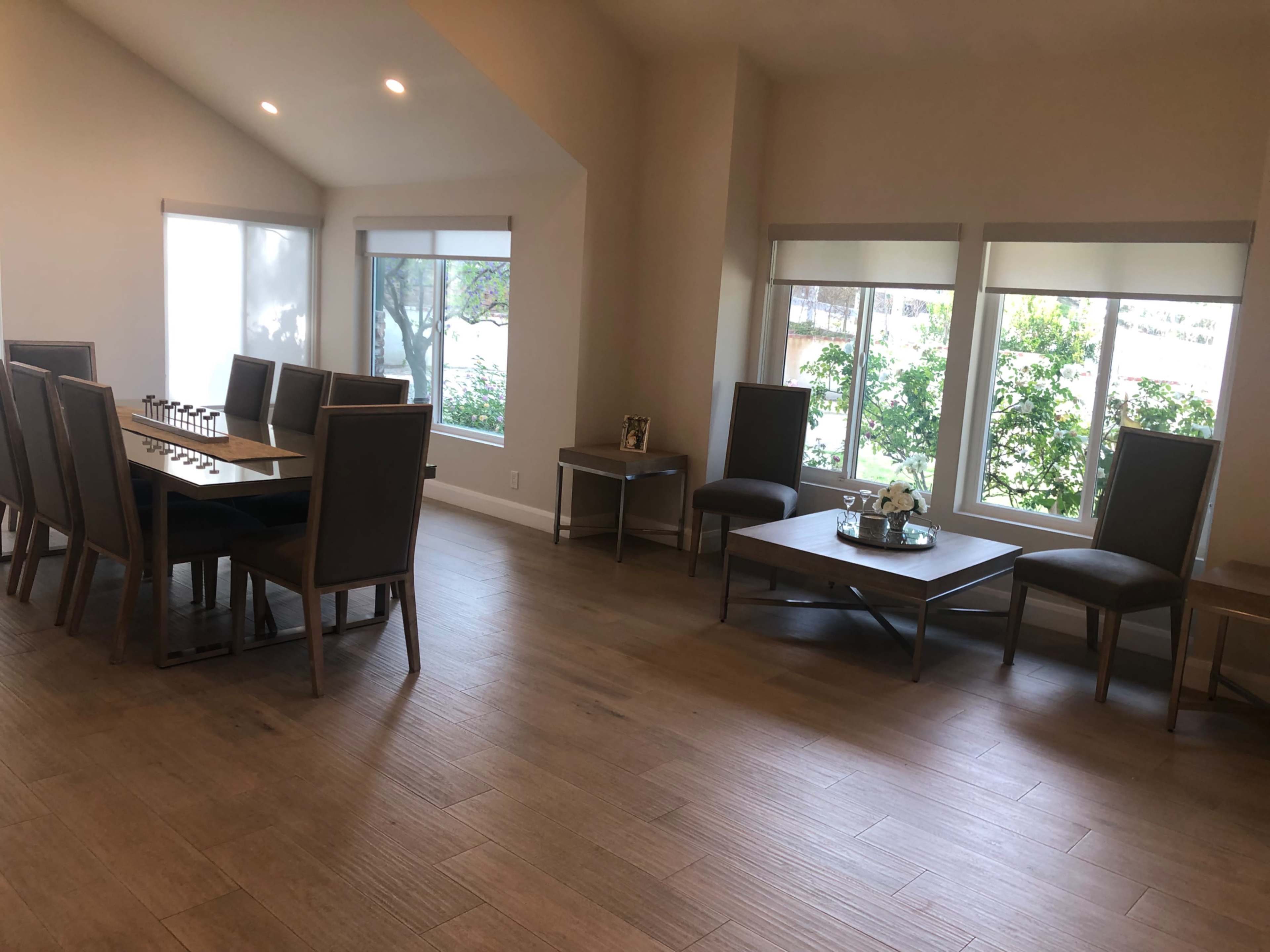 A spacious room features a long table with chairs on one side and several smaller tables with chairs on the other side, all illuminated by natural light from large windows.