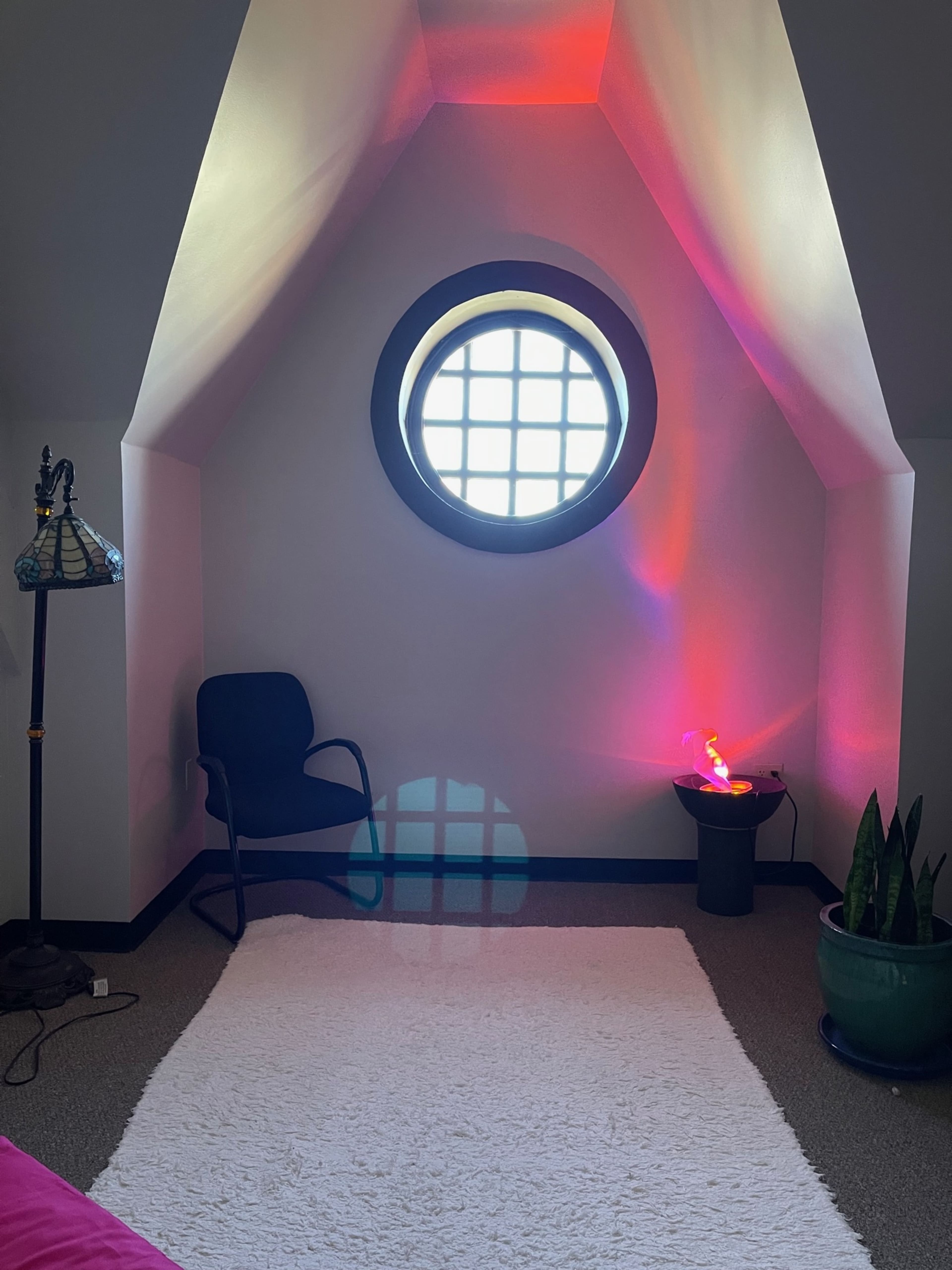 A corner of a room features a round window, a small plant, a lamp, and a chair on a white rug.