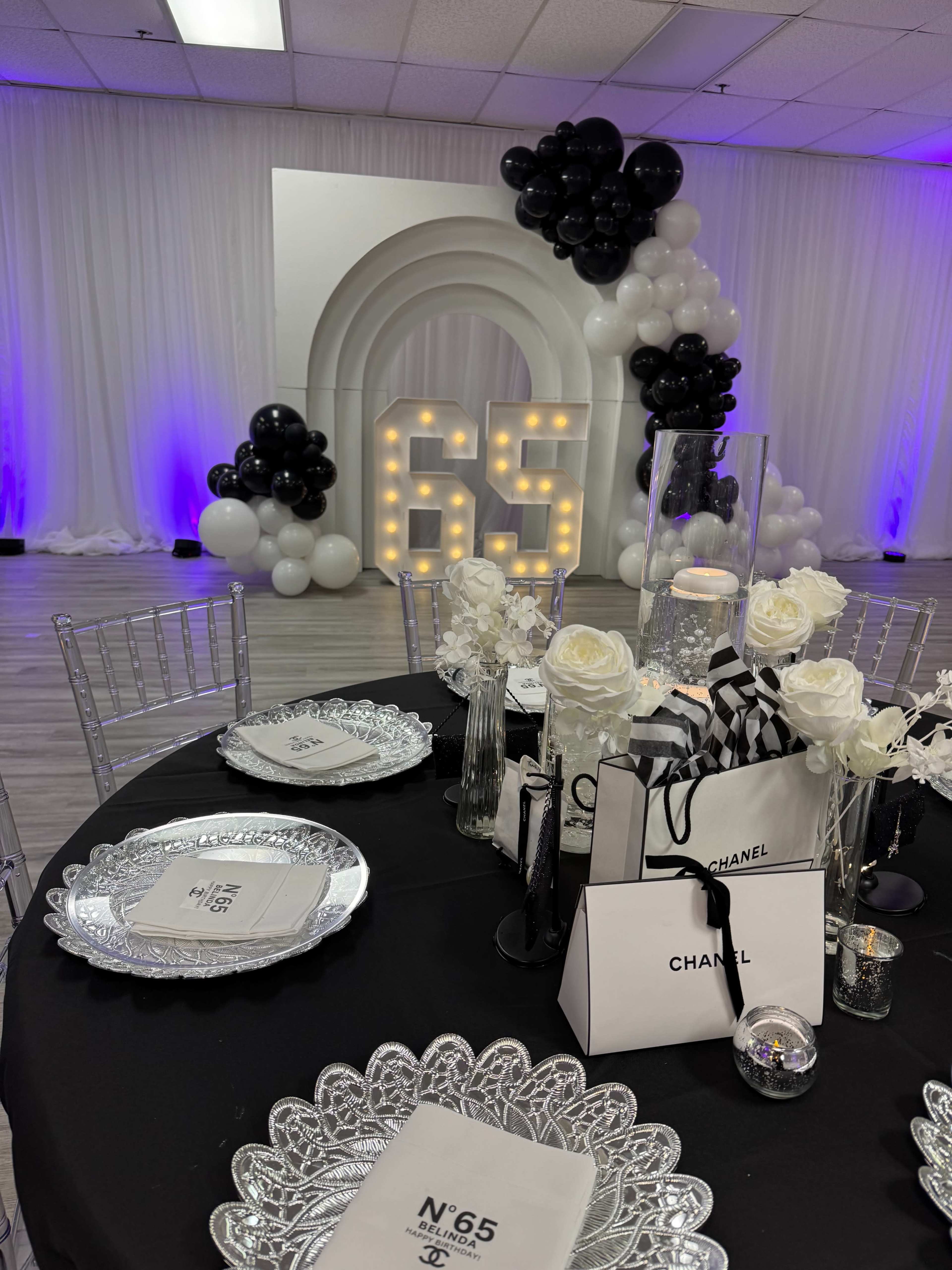 The image depicts a decorated event space featuring a table set with silver plates and black and white accents, along with a large display of balloons and illuminated numbers reading "65" in the background.