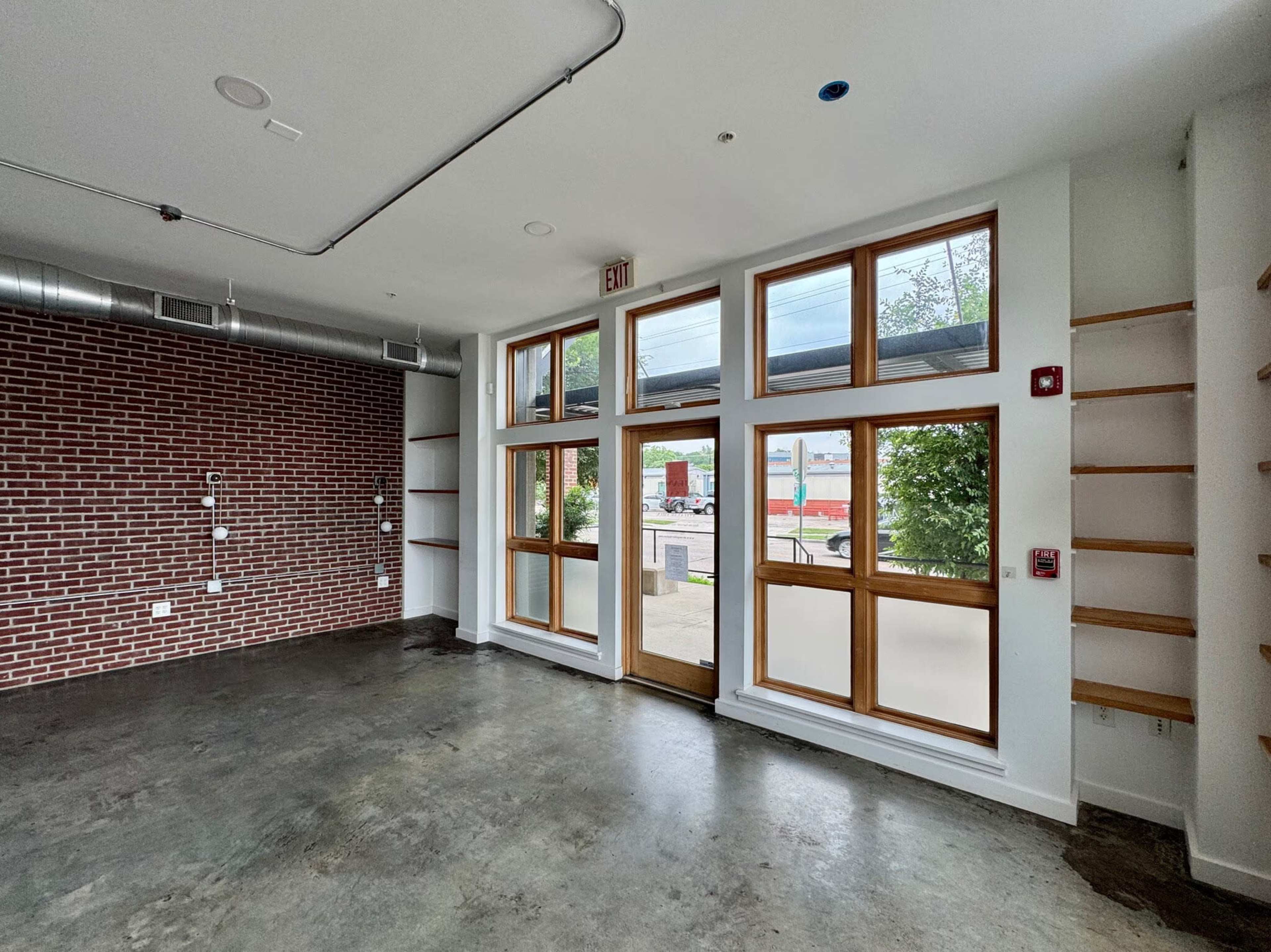 The image shows an empty room featuring a brick wall, large windows with wooden frames, and shelving along one side.