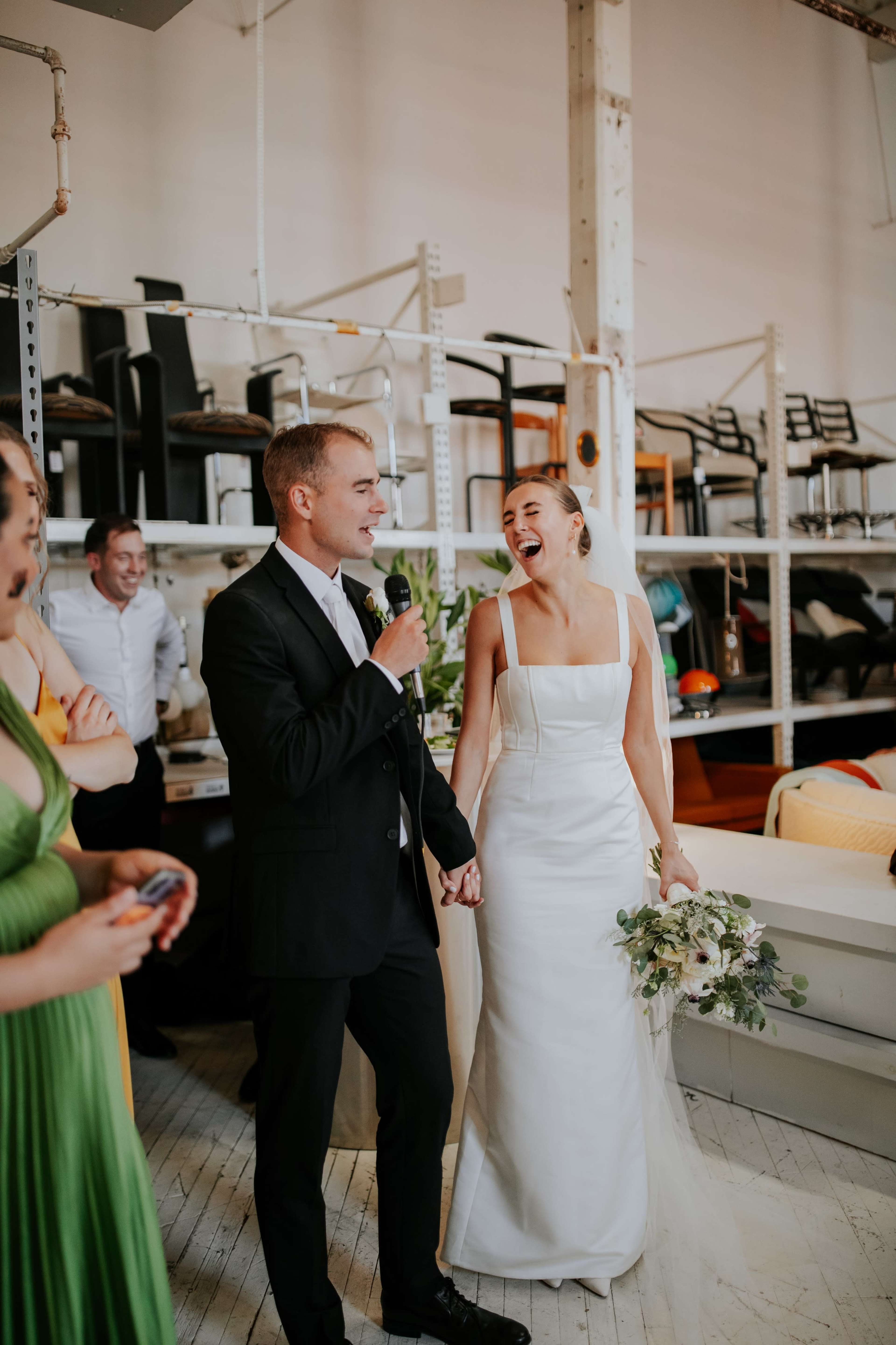 A man in a suit speaks into a microphone while holding hands with a smiling woman in a white wedding dress, surrounded by guests in a bright, industrial-style venue.
