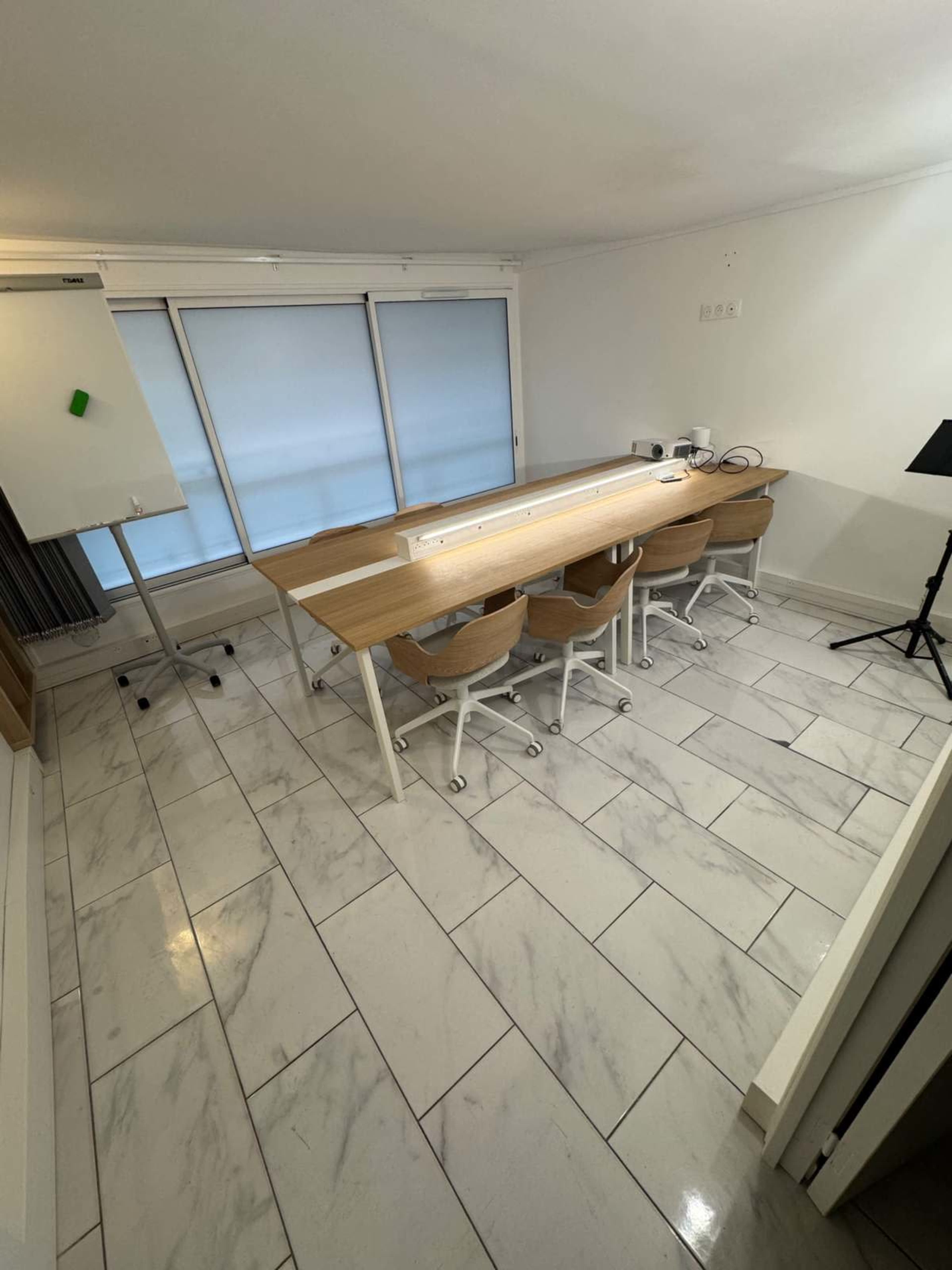 The image shows a modern meeting room with a long table, several chairs, a whiteboard, and large windows partially covered by blinds.