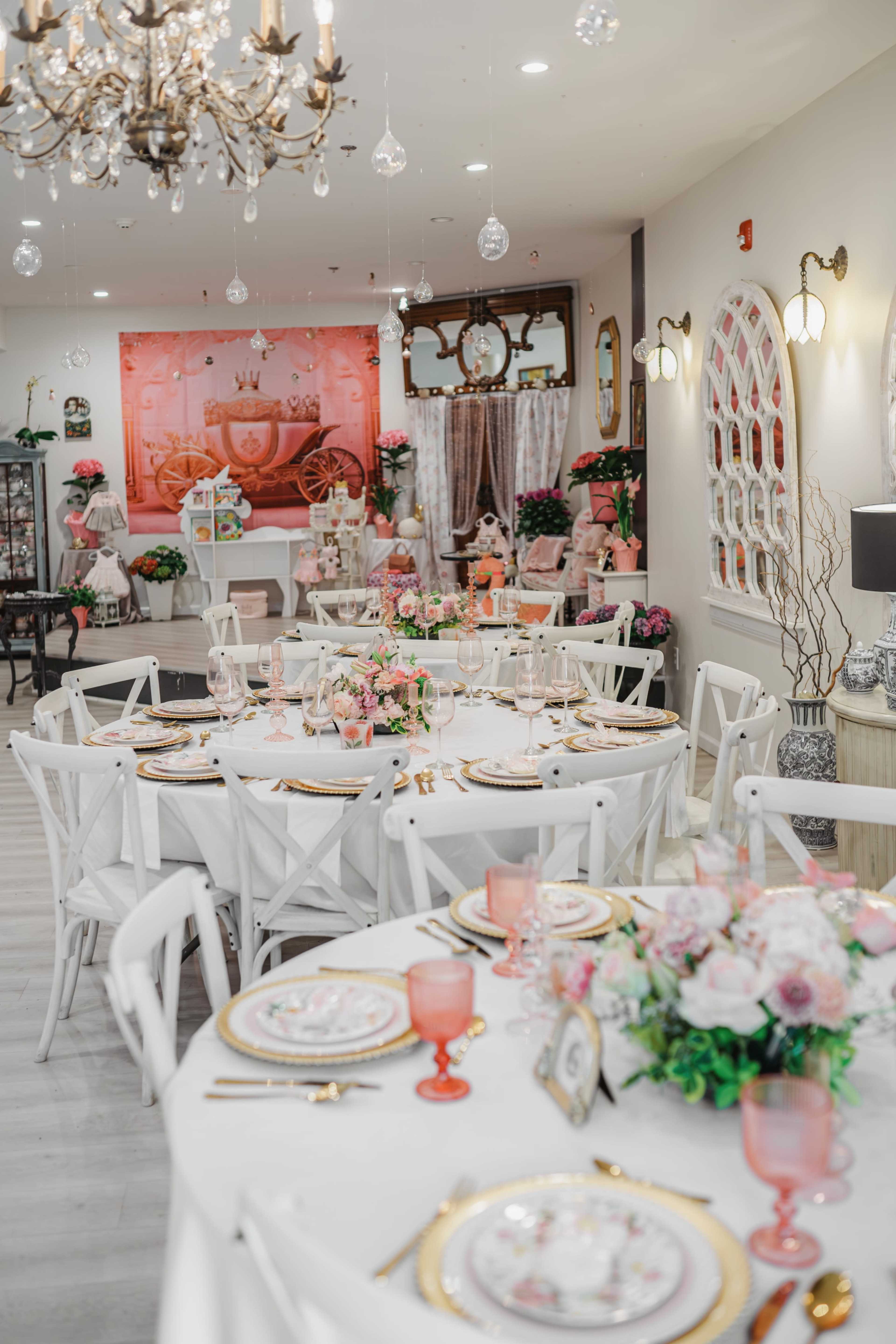 A beautifully arranged dining area features round tables set with floral centerpieces, elegant dishware, and a chandelier overhead in a bright, decorated space.