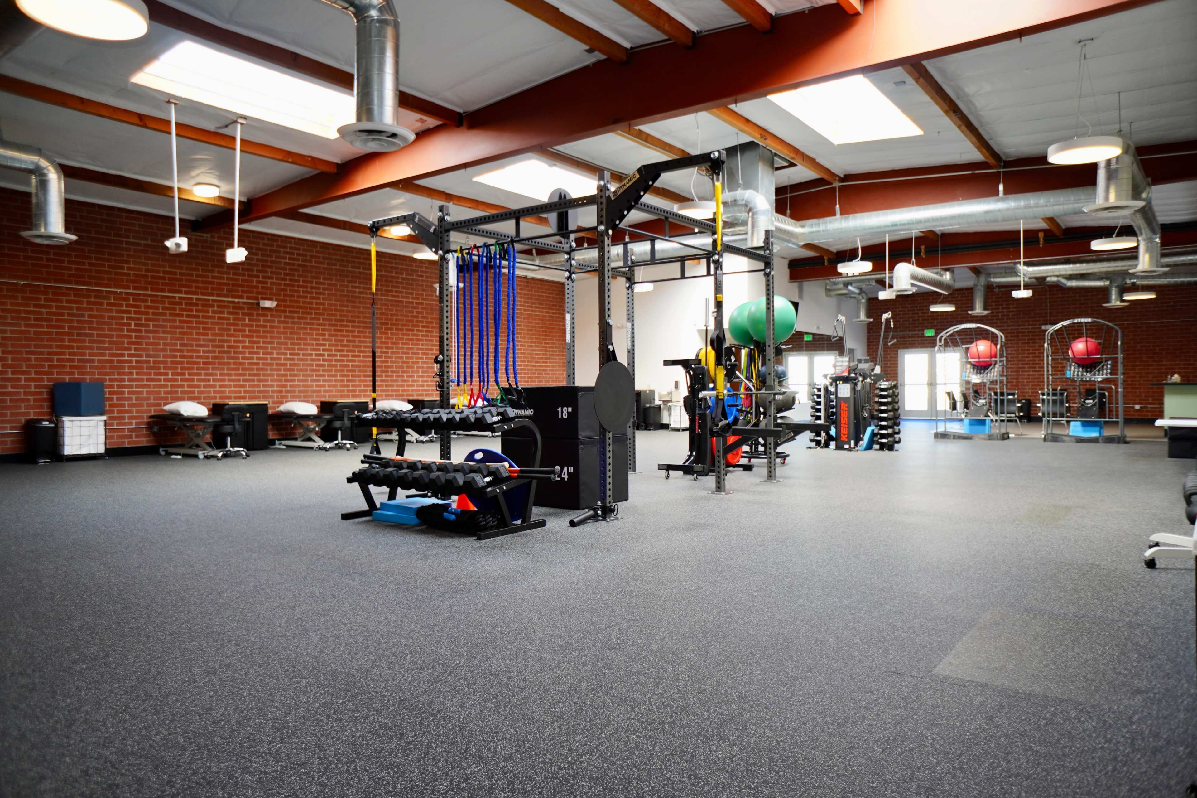 A spacious gym interior equipped with various weightlifting and exercise equipment, including free weights and resistance bands, under a high ceiling with exposed beams.