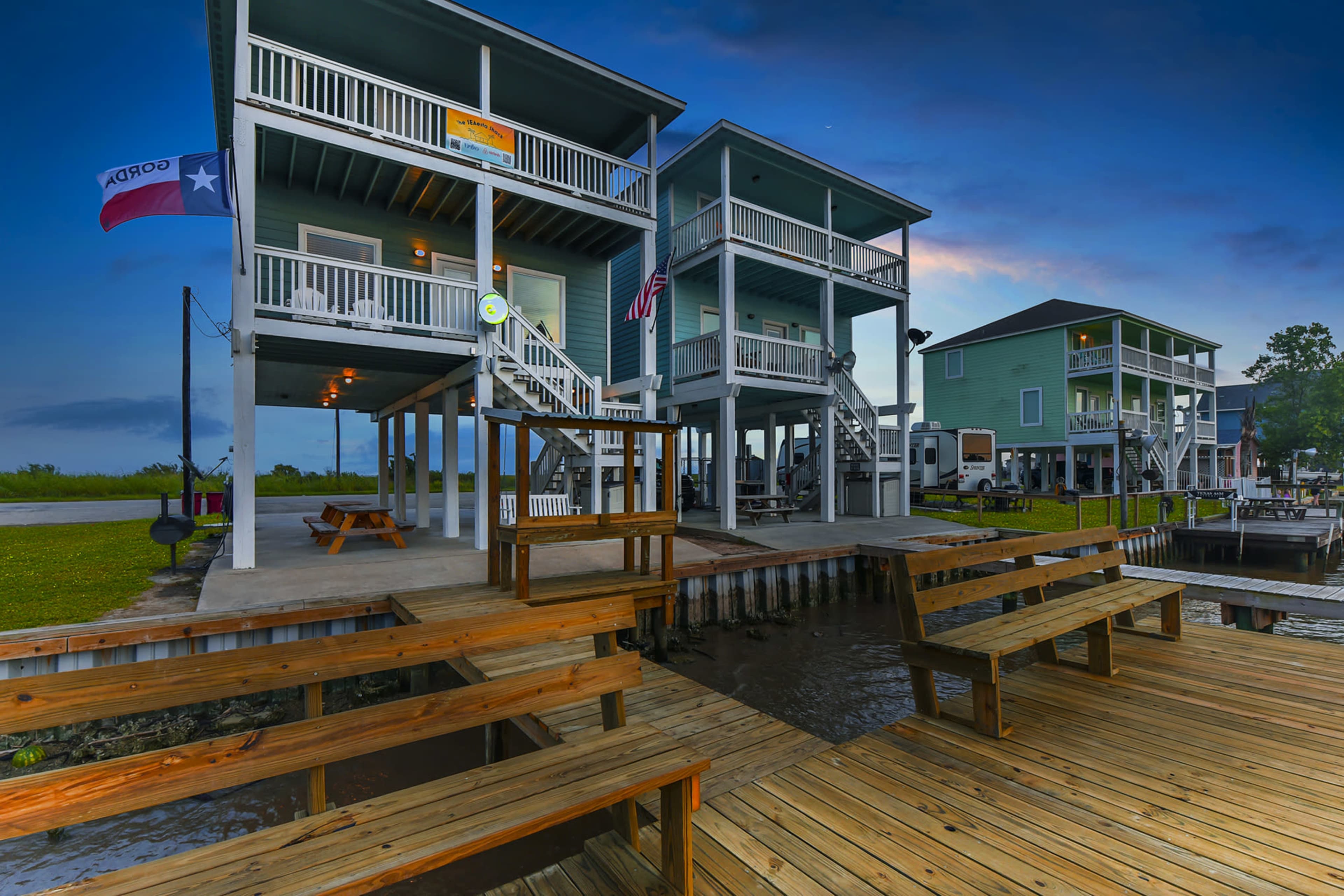 The image features several elevated houses with porches, situated near a waterway, along with a wooden dock and picnic tables.