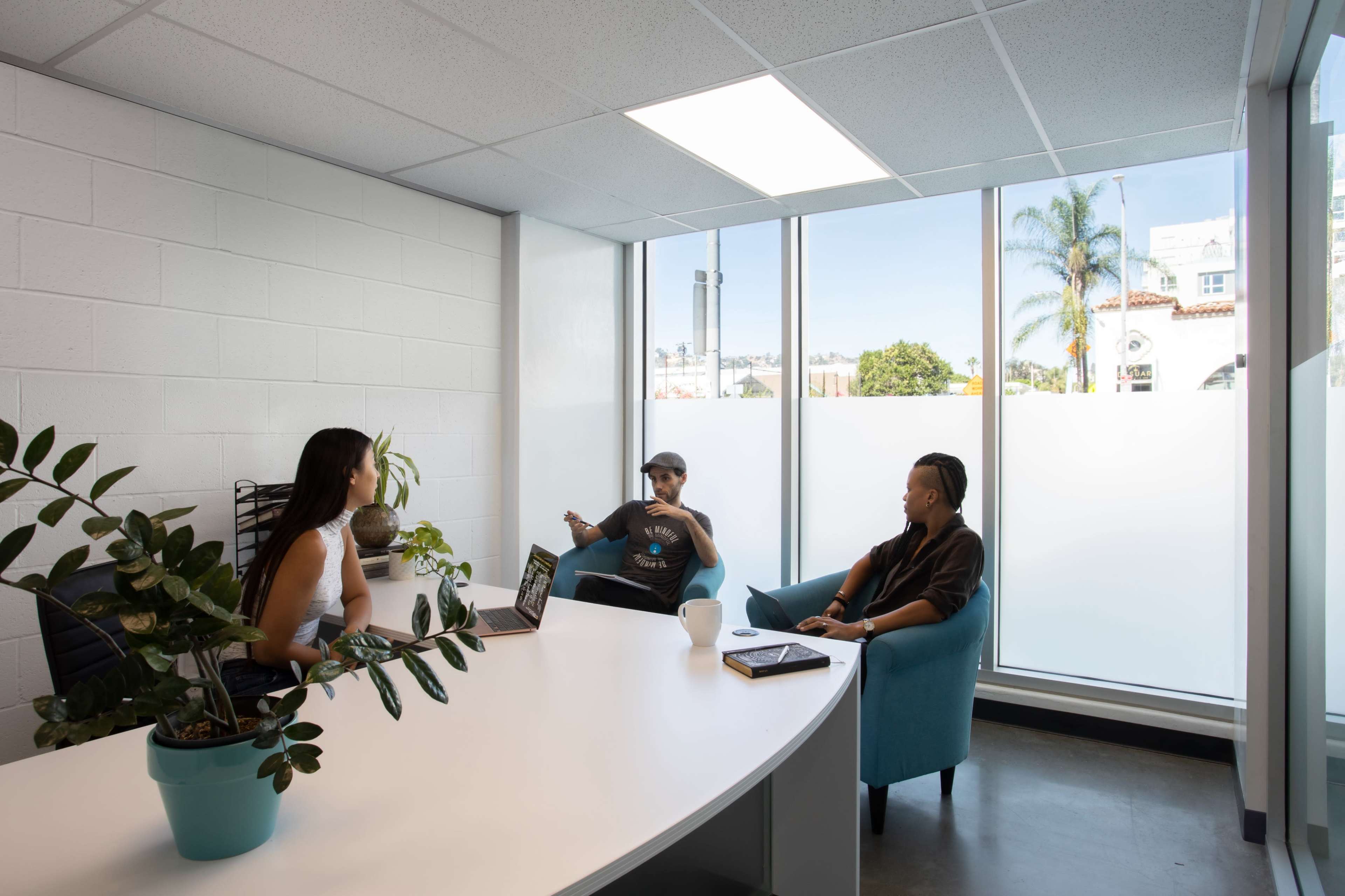 Three people are seated in a bright office space with large windows, discussing around a white table.