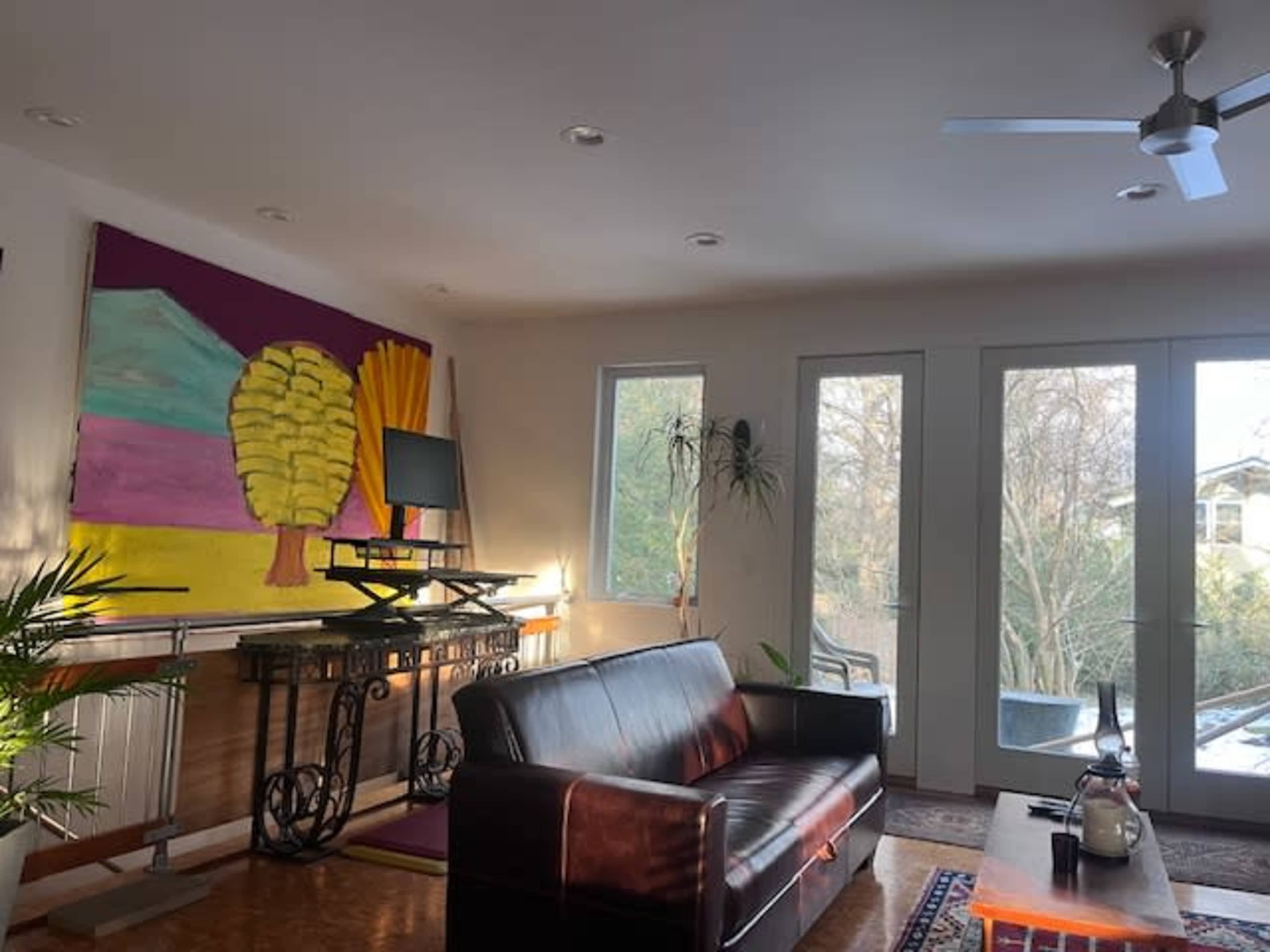 The image shows a modern living room with a dark leather sofa, a large colorful painting on the wall, and glass doors opening to a view outside.