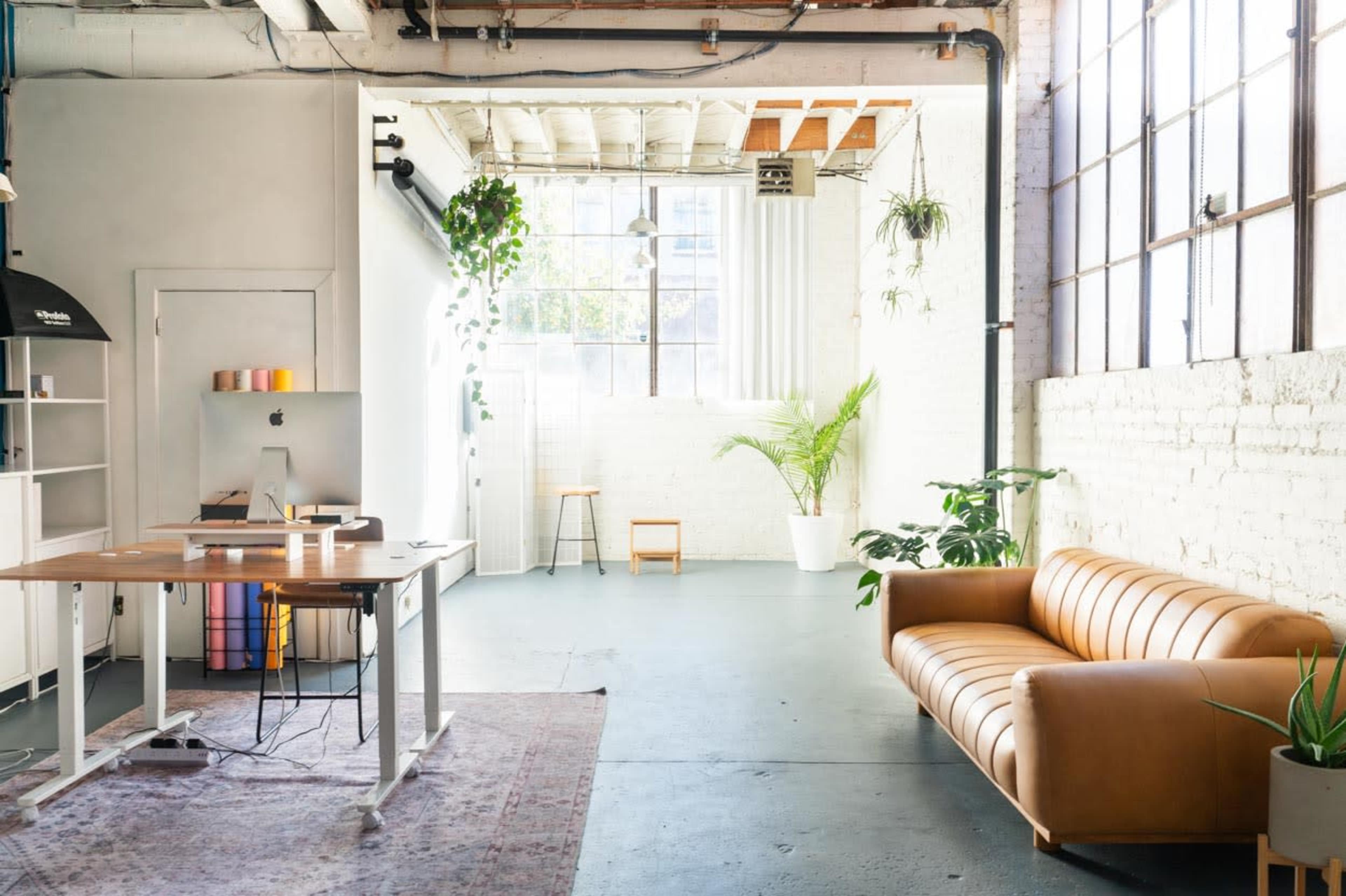 A bright, open workspace featuring a wooden desk with a computer, a brown leather couch, and plants in several areas against white walls and large windows.