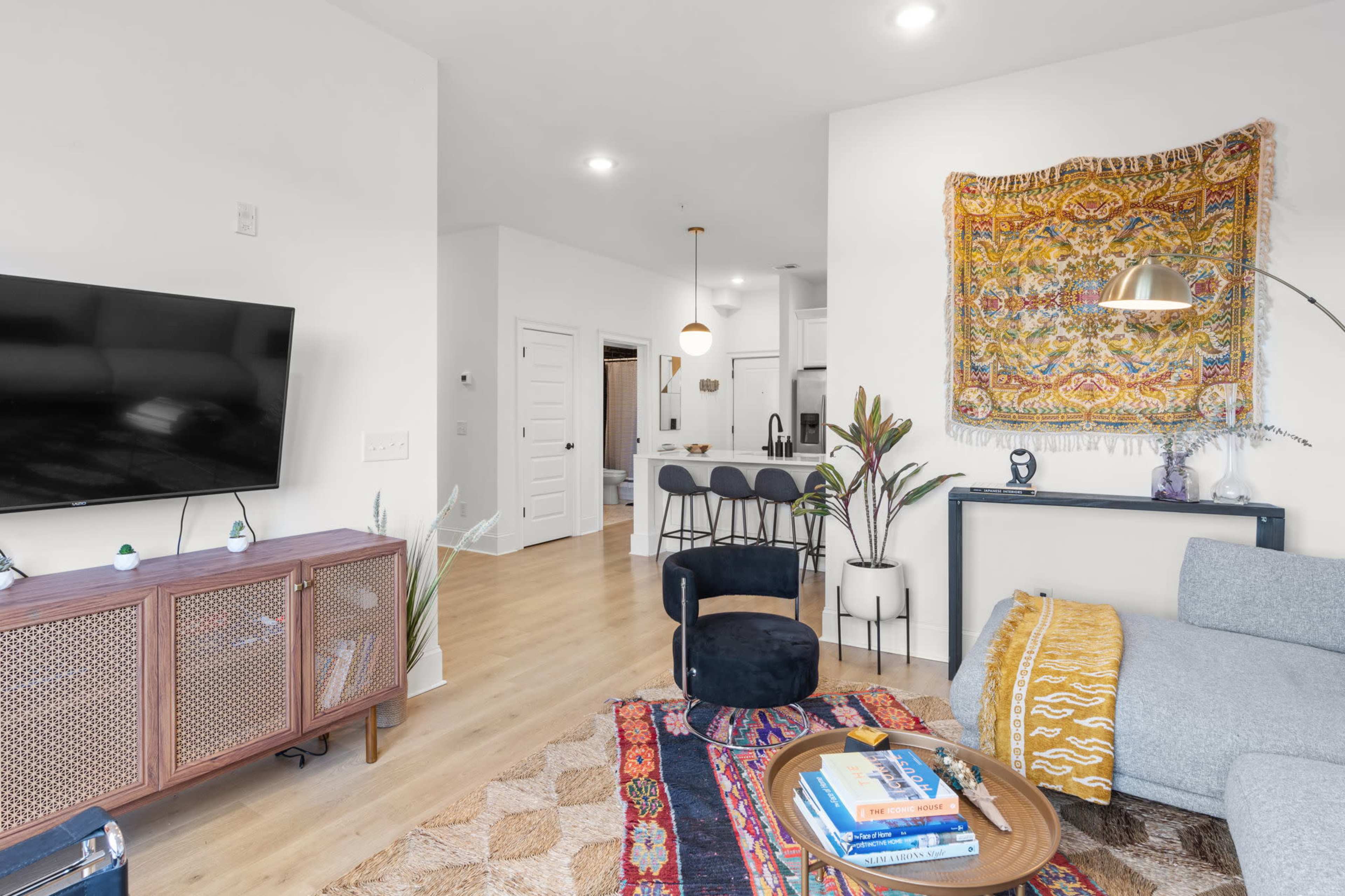 The image shows a modern living room with a television mounted on the wall, a patterned rug on the floor, and a cozy seating area, including a gray sofa and a round coffee table.