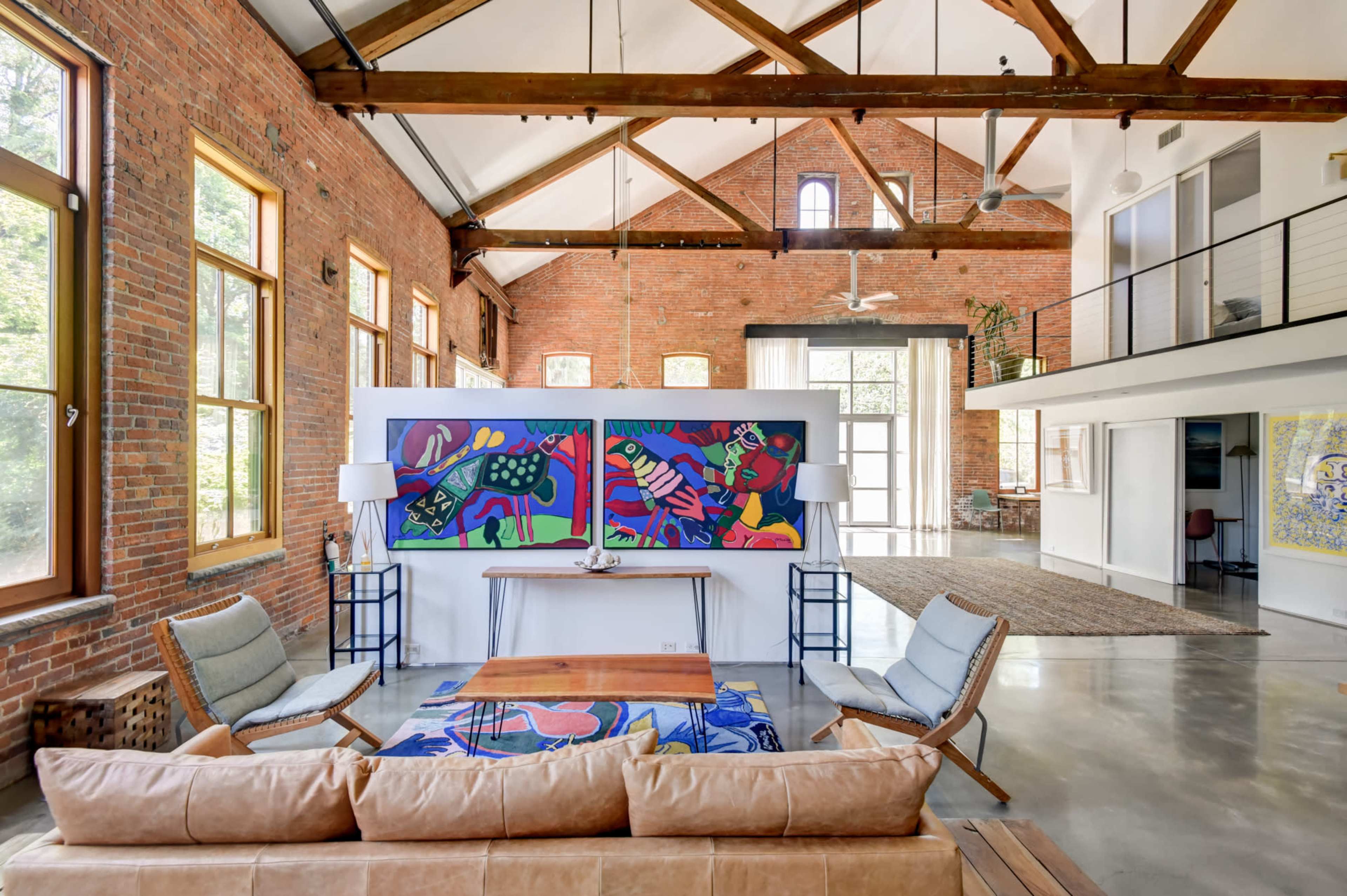 A spacious interior features exposed brick walls, high ceilings with wooden beams, and large windows that allow natural light to illuminate the room, which includes a sofa, artwork, and a table.