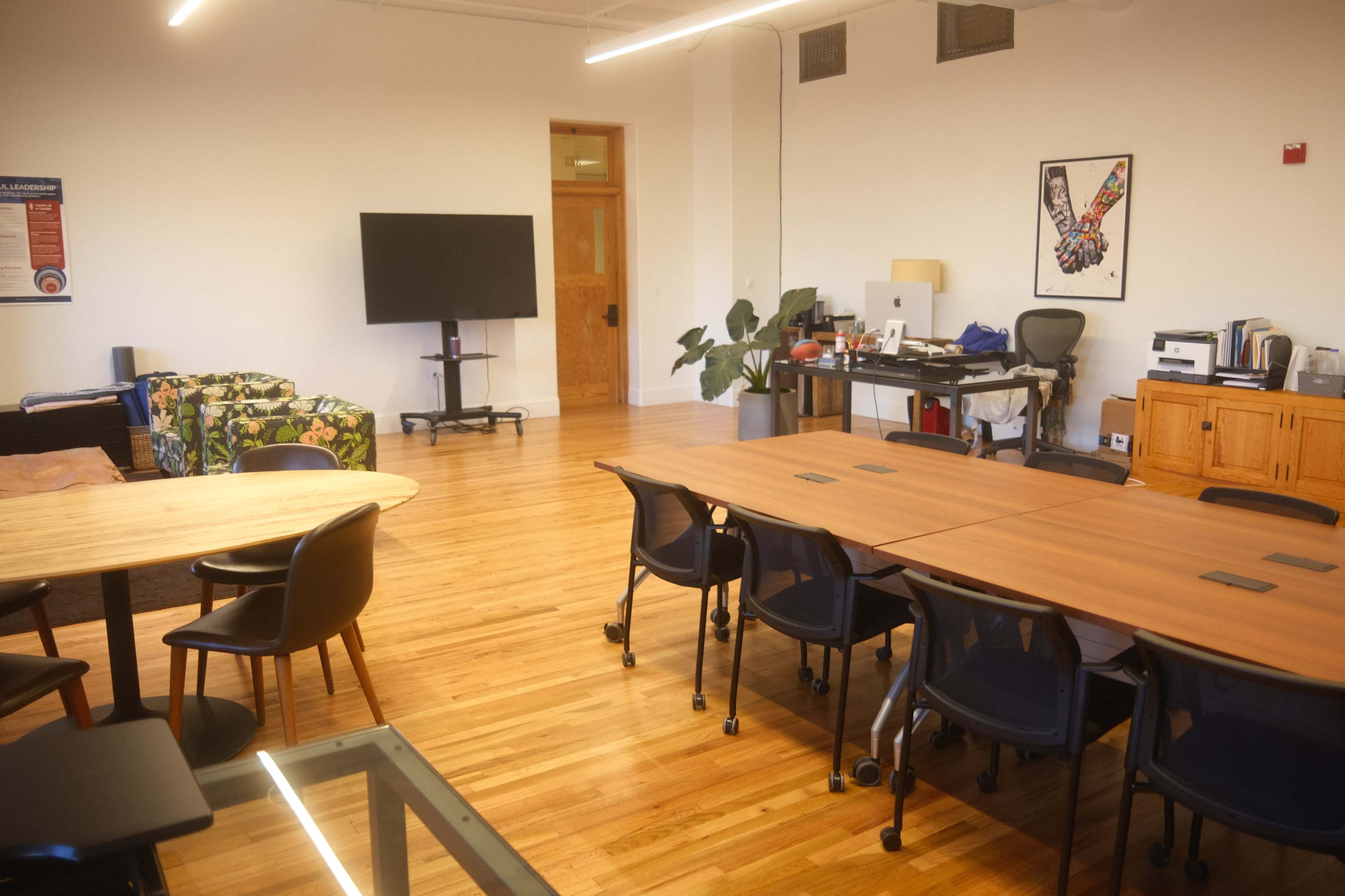 The image shows a spacious meeting room with several tables set up, a TV on a stand, a desk area with a computer, and a colorful couch in the corner.