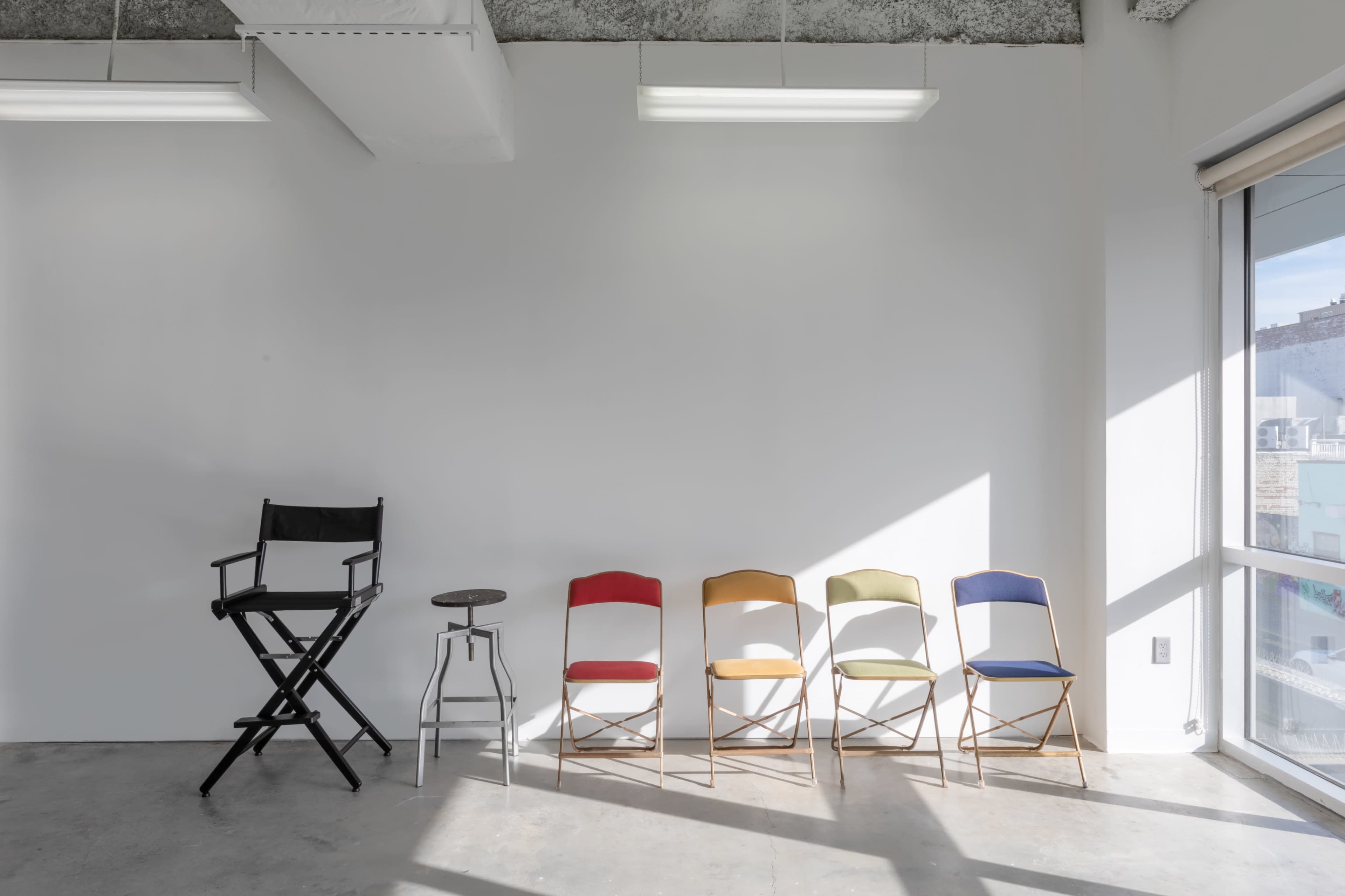 A black director's chair is positioned next to a metal stool and five colorful folding chairs against a white wall in a well-lit room.