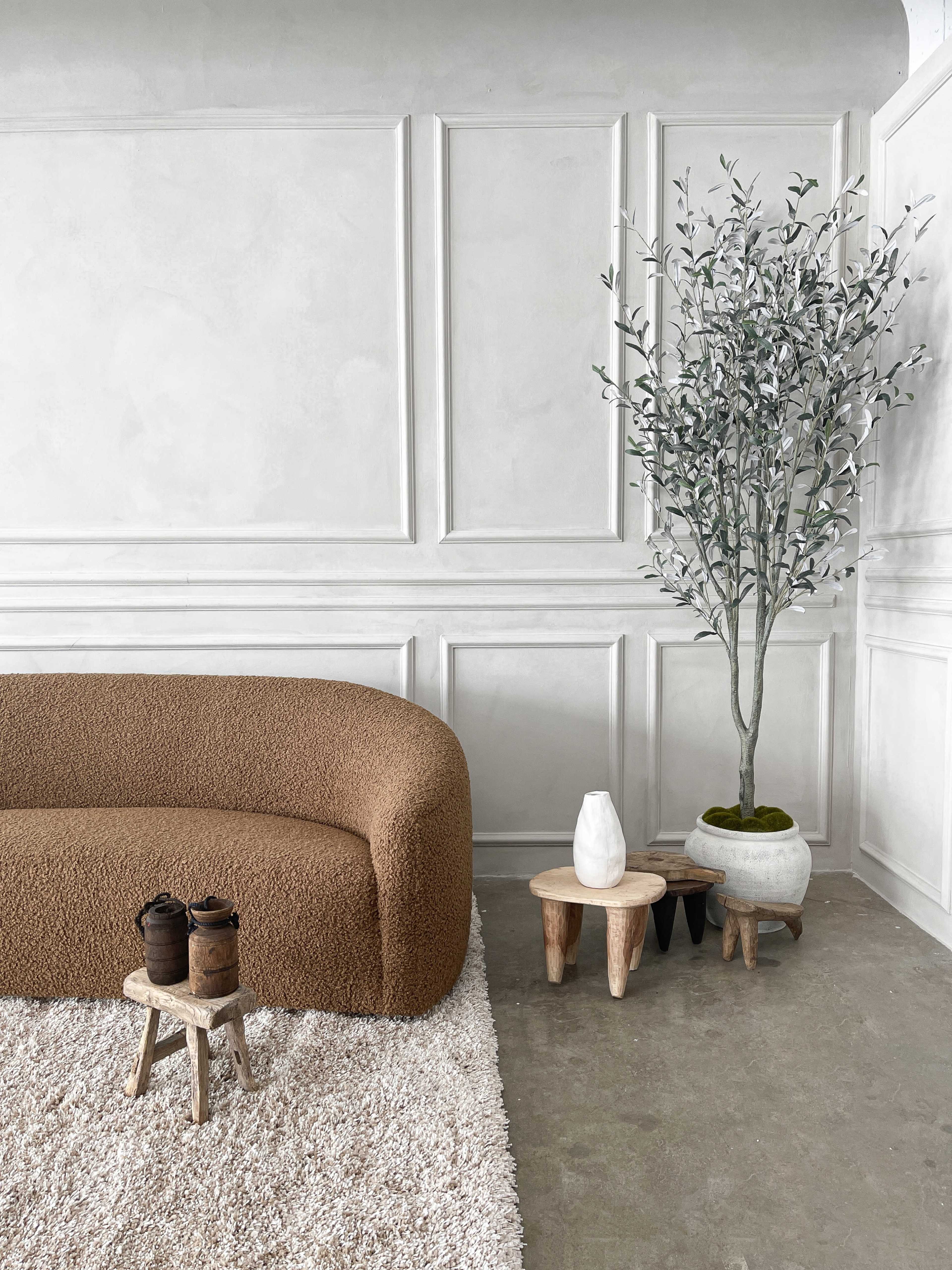 A modern living space featuring a brown curved sofa, two small wooden stools, a white decorative vase, and a potted tree against a textured white wall.