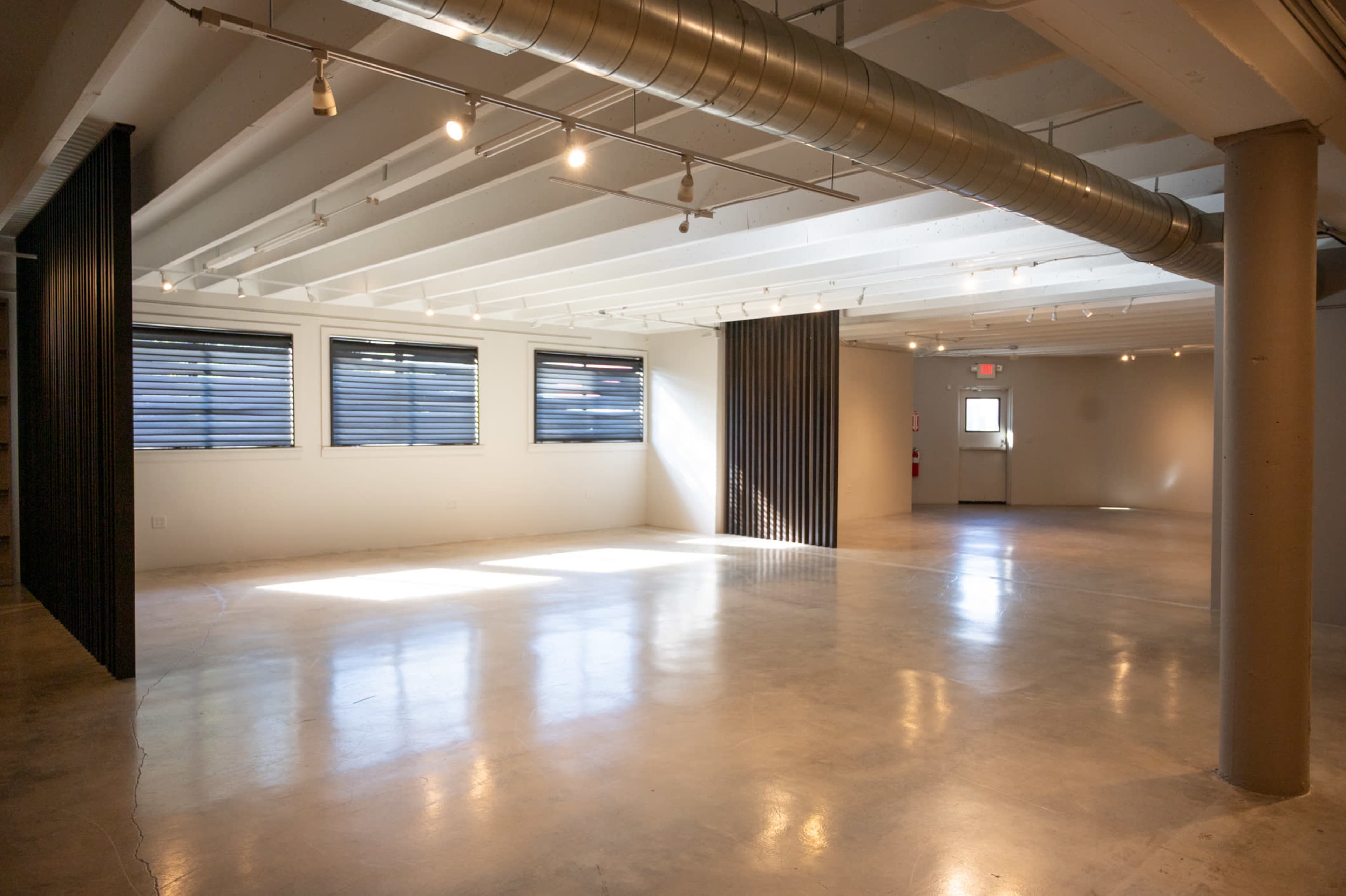 The image shows a spacious, empty room with polished concrete flooring, large windows, and exposed ventilation pipes.