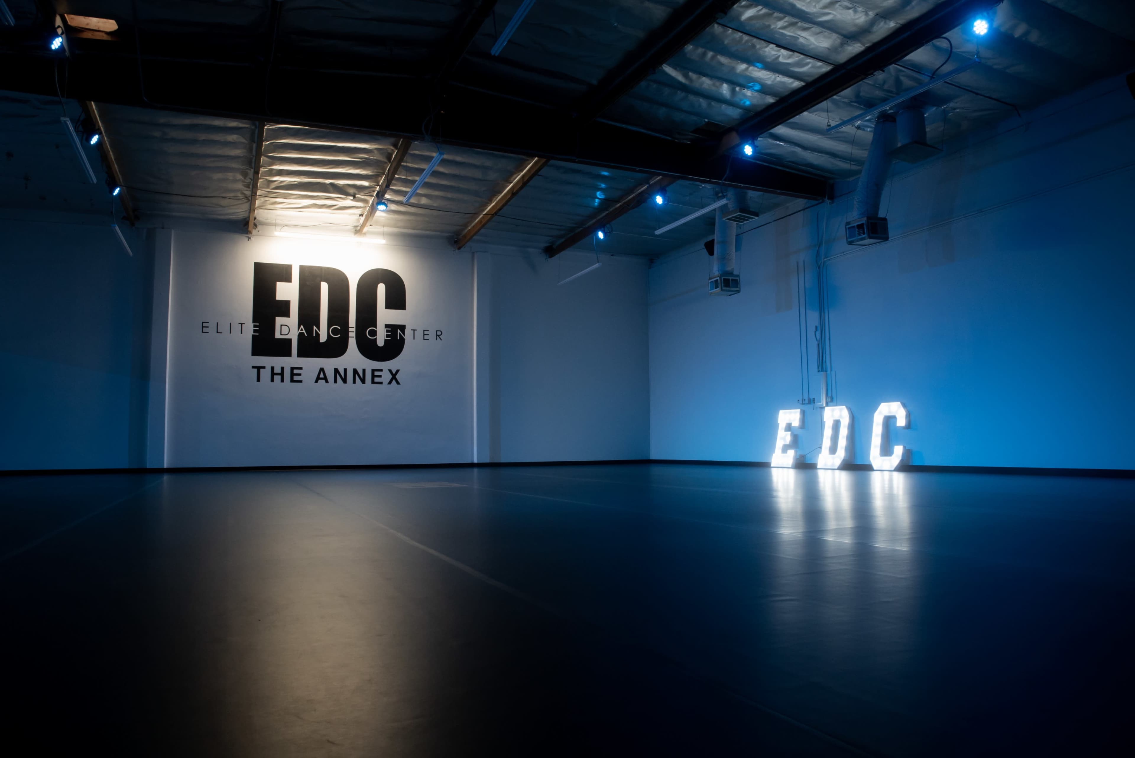 An empty dance studio with a wall displaying the logo "EDC Elite Dance Center" and illuminated letters spelling "EDC" on the floor.