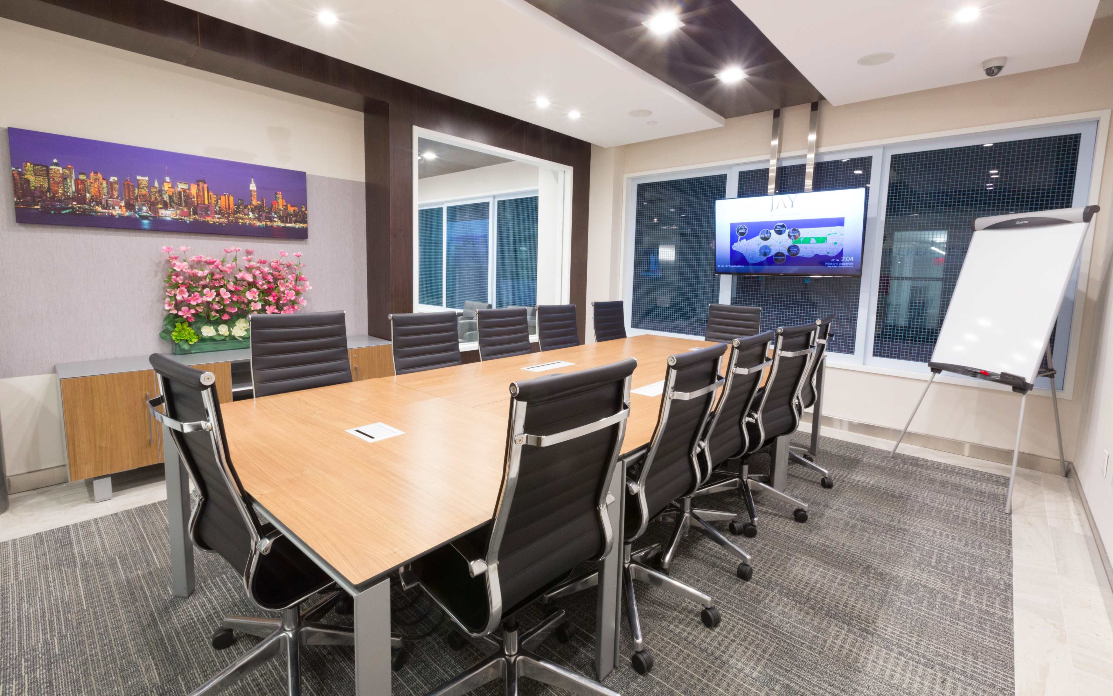 The image shows a modern conference room with a long wooden table, black chairs, and a large screen on the wall displaying a presentation.