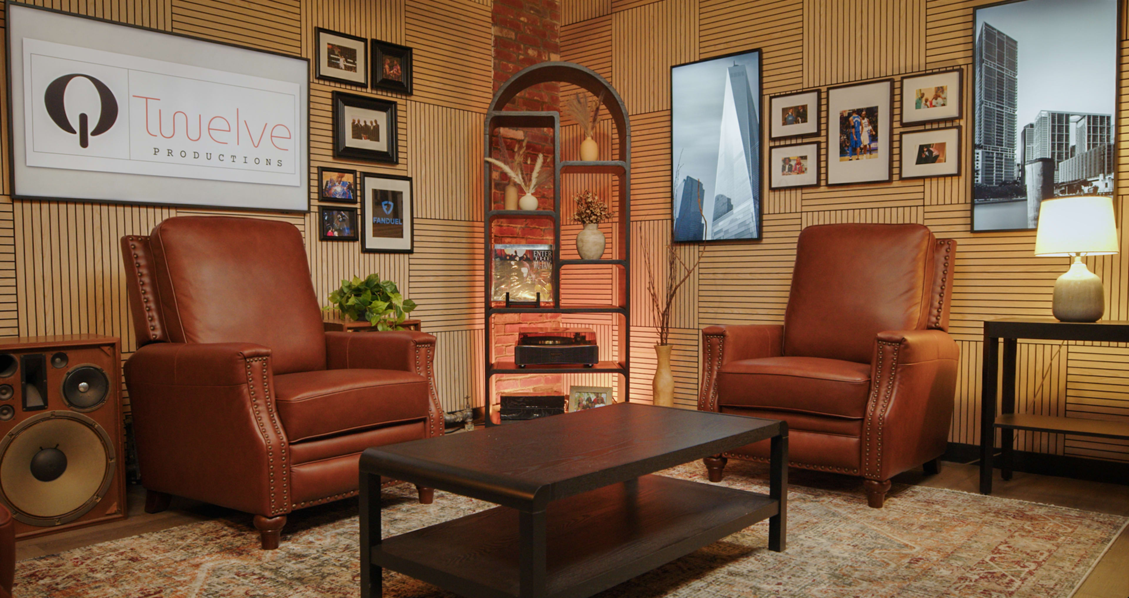 The image shows a stylish interior featuring two brown leather armchairs facing a wooden coffee table, with framed photos and screens mounted on the walls amid wooden paneling.