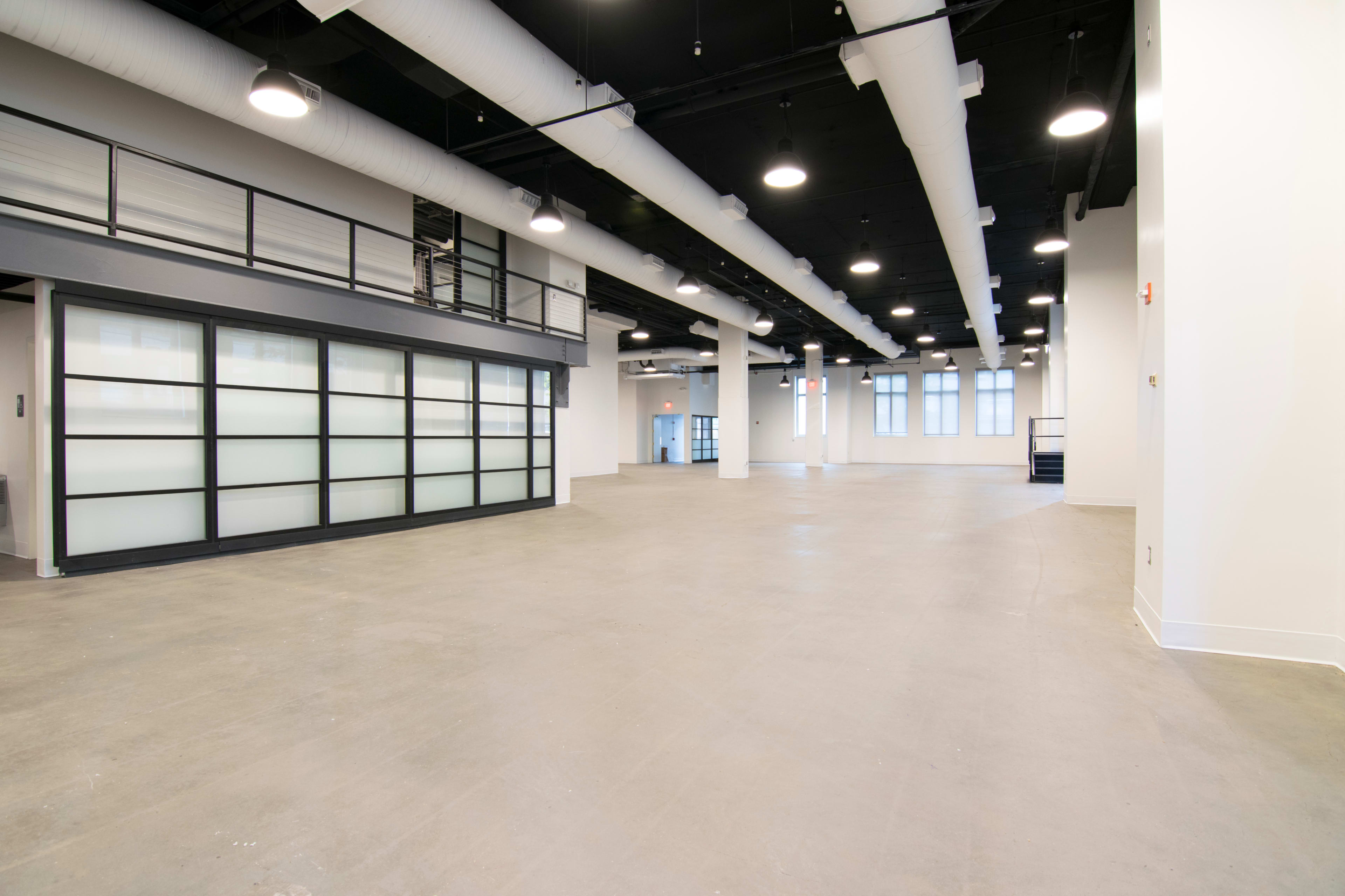 A spacious, empty room with high ceilings, industrial lighting, and a glass wall dividing an adjacent area.