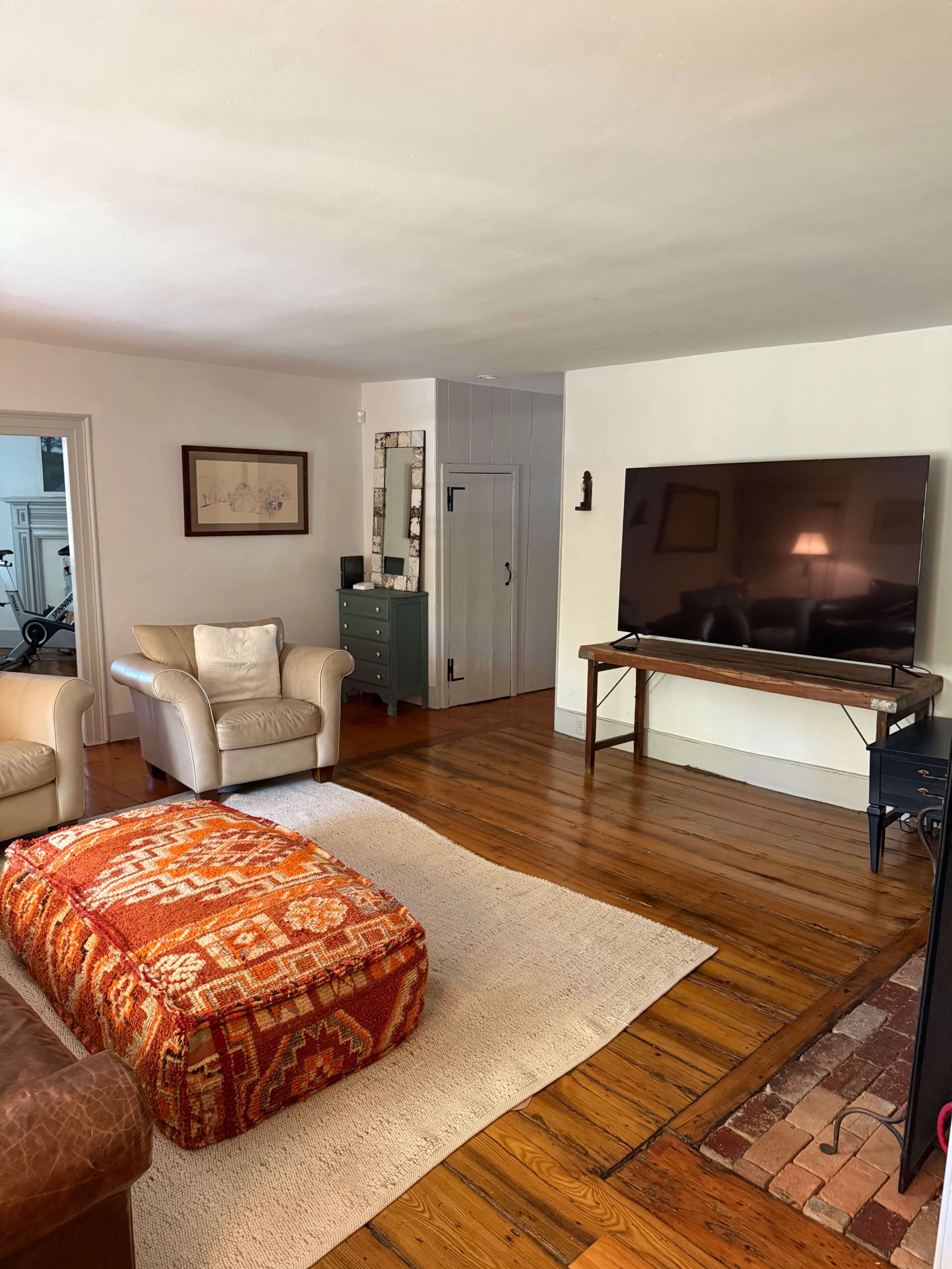 A cozy living room features a large television on a wooden stand, a plush ottoman with a patterned design, and two armchairs on a wooden floor.