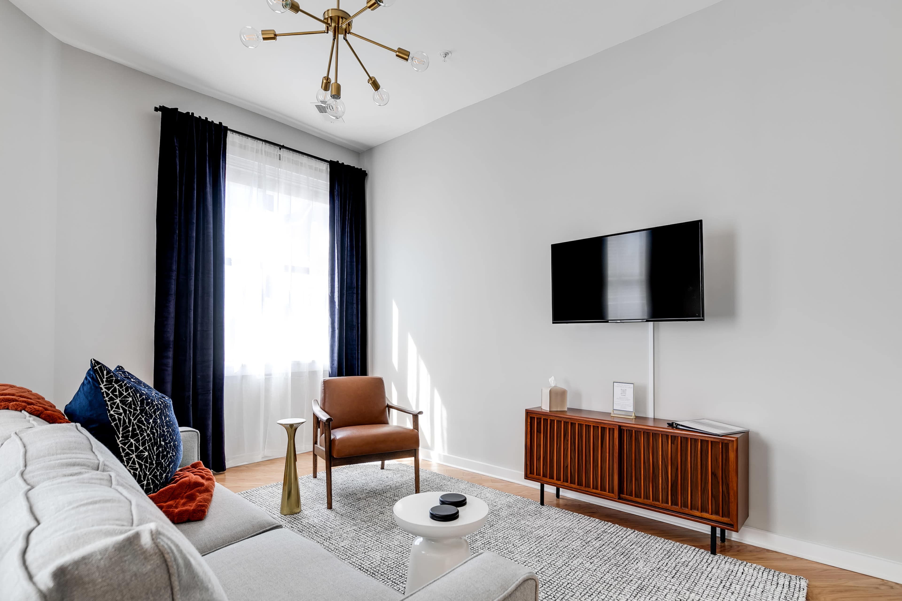 A modern living room with a gray sofa, a wooden cabinet, a television on the wall, and a brown armchair near a window with sheer curtains.