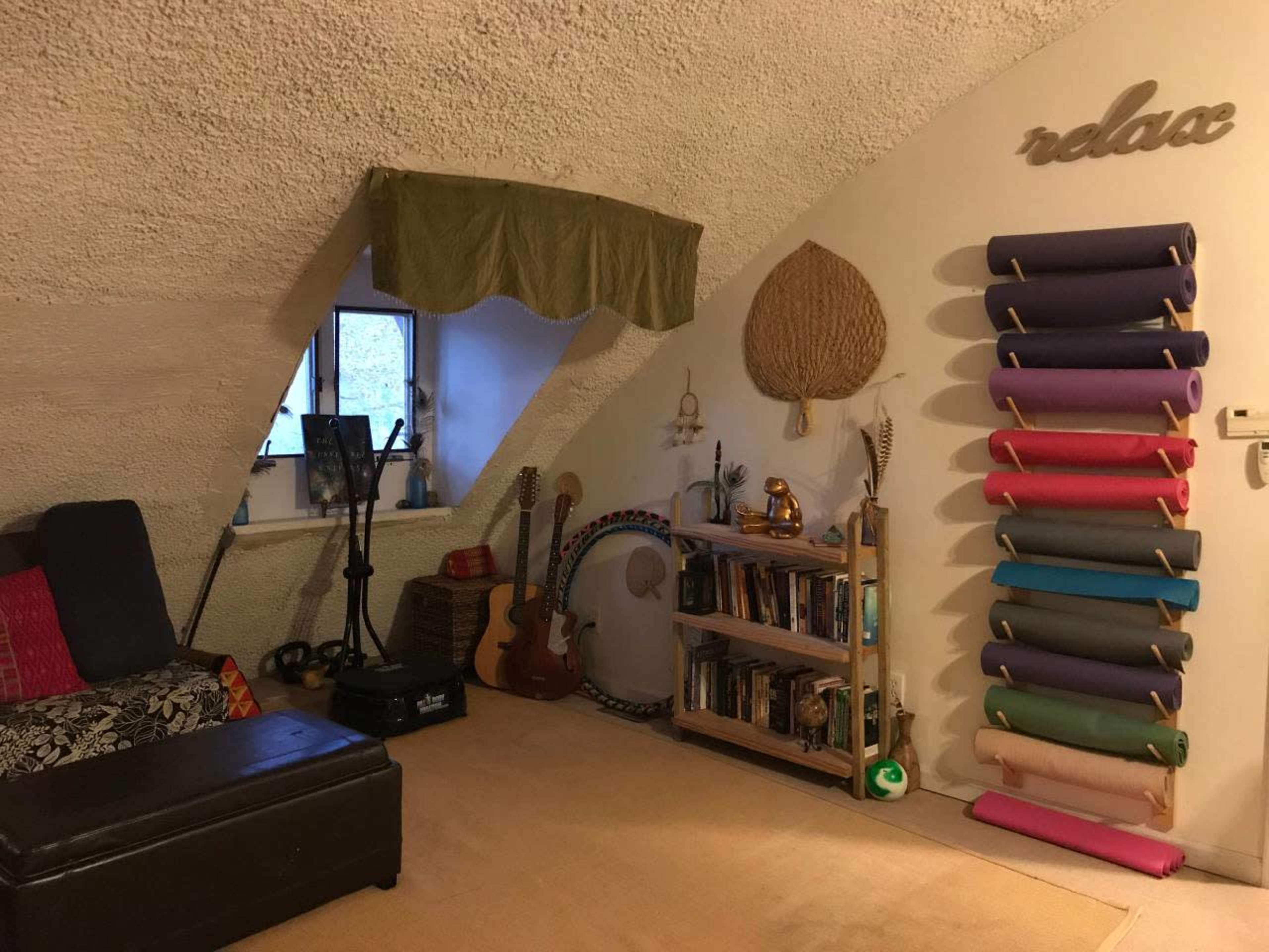 The image shows a cozy, curved room with a dark sofa, a bookshelf filled with books, a guitar, and colorful rolls of fabric arranged on the wall.