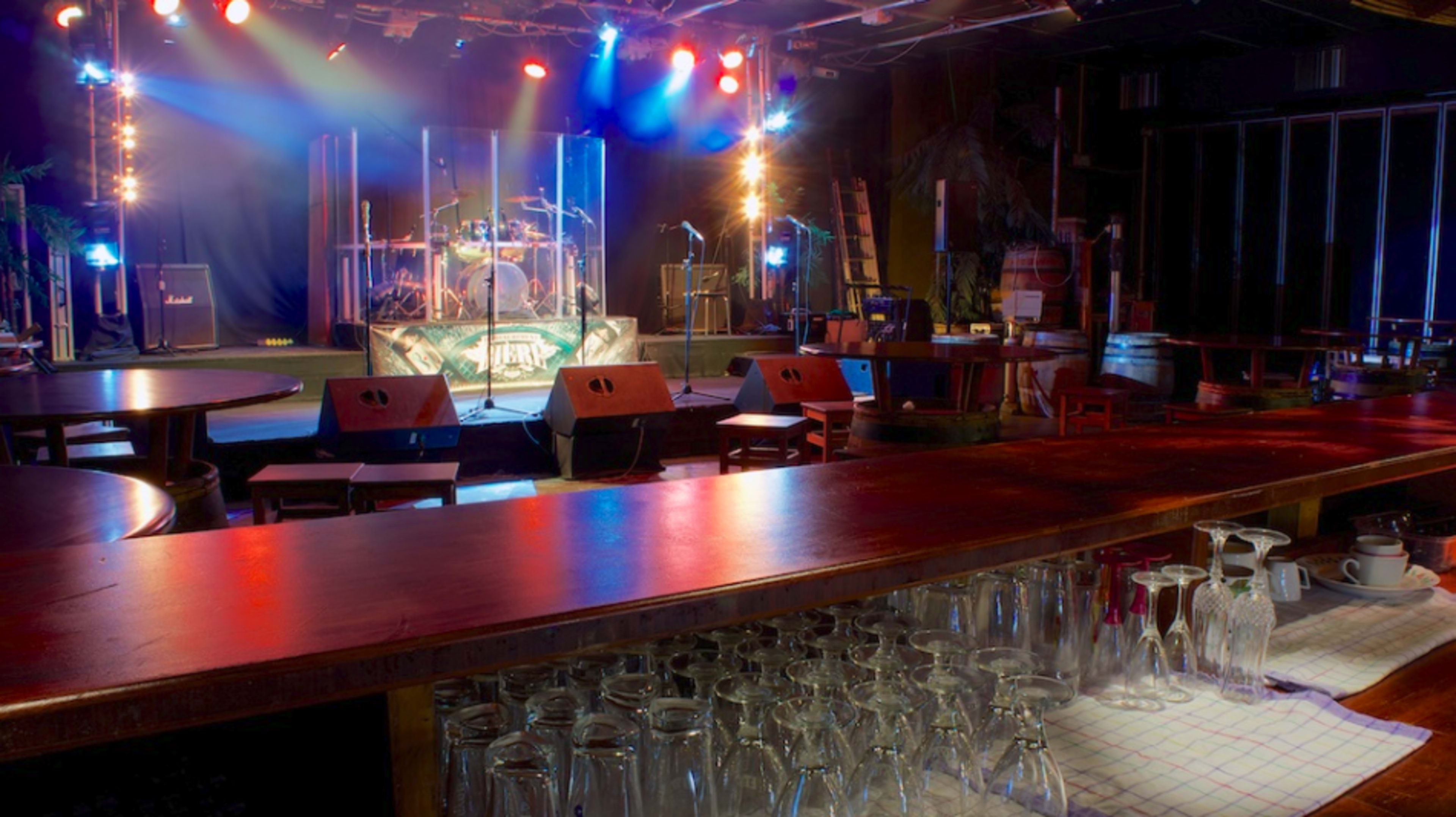 A music venue features a stage with instruments and lighting, flanked by empty tables and stacked glasses in the foreground.