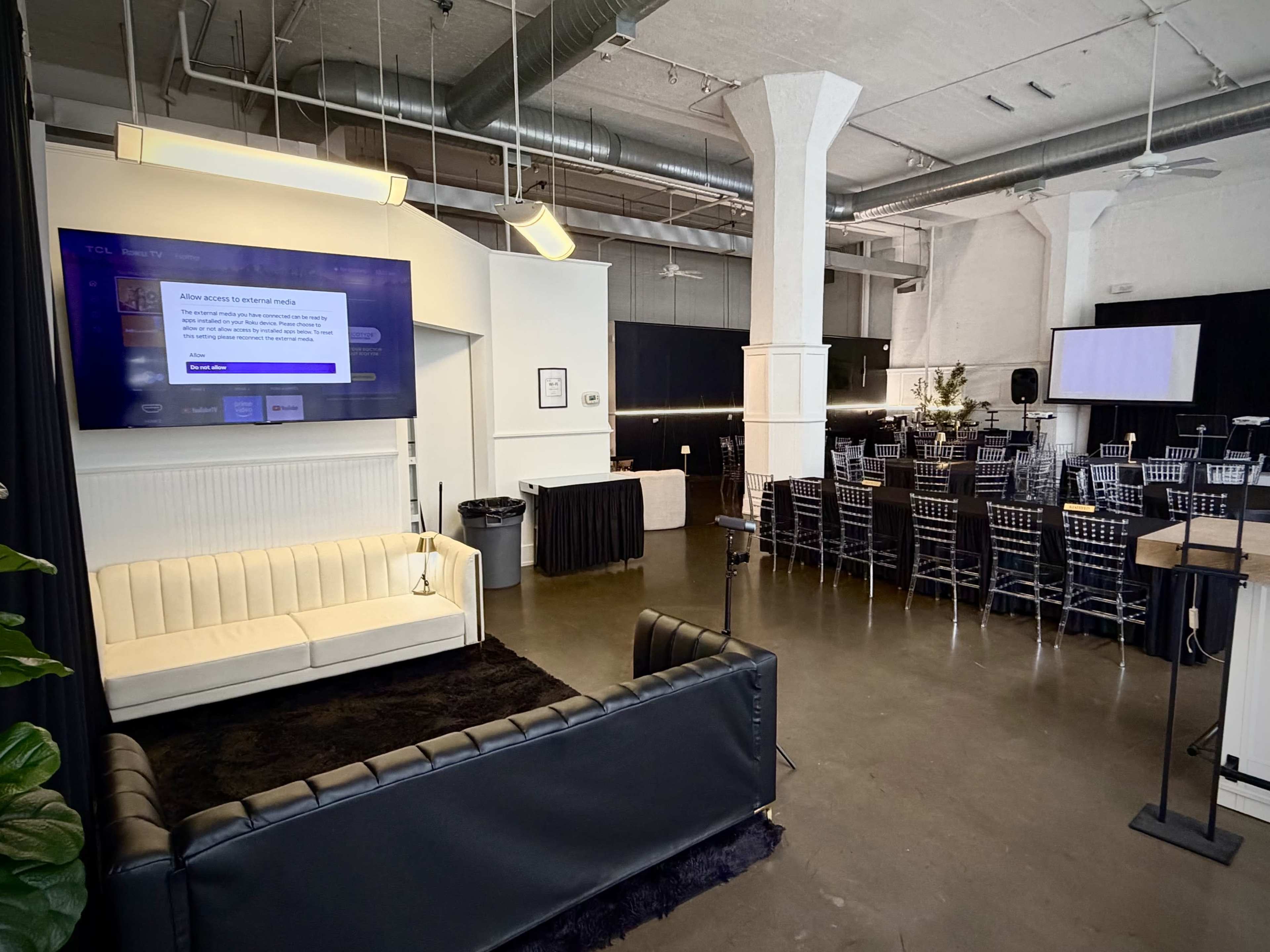 The image shows a spacious indoor venue featuring a seating area with a couch, a large screen displaying a message, and rows of tables and chairs set up for an event.