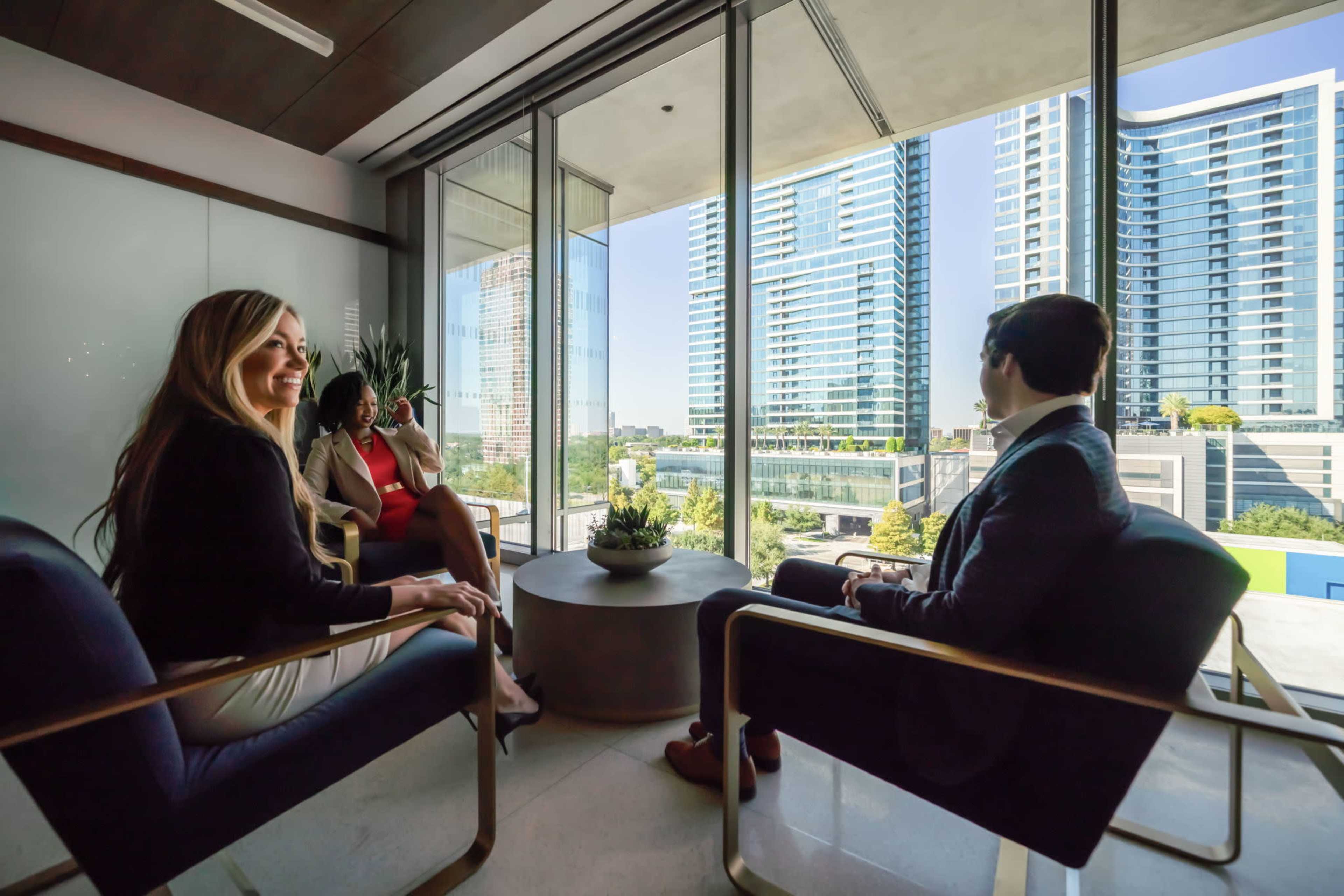 Three professionals are seated in a modern conference room with glass walls, overlooking a cityscape featuring high-rise buildings.