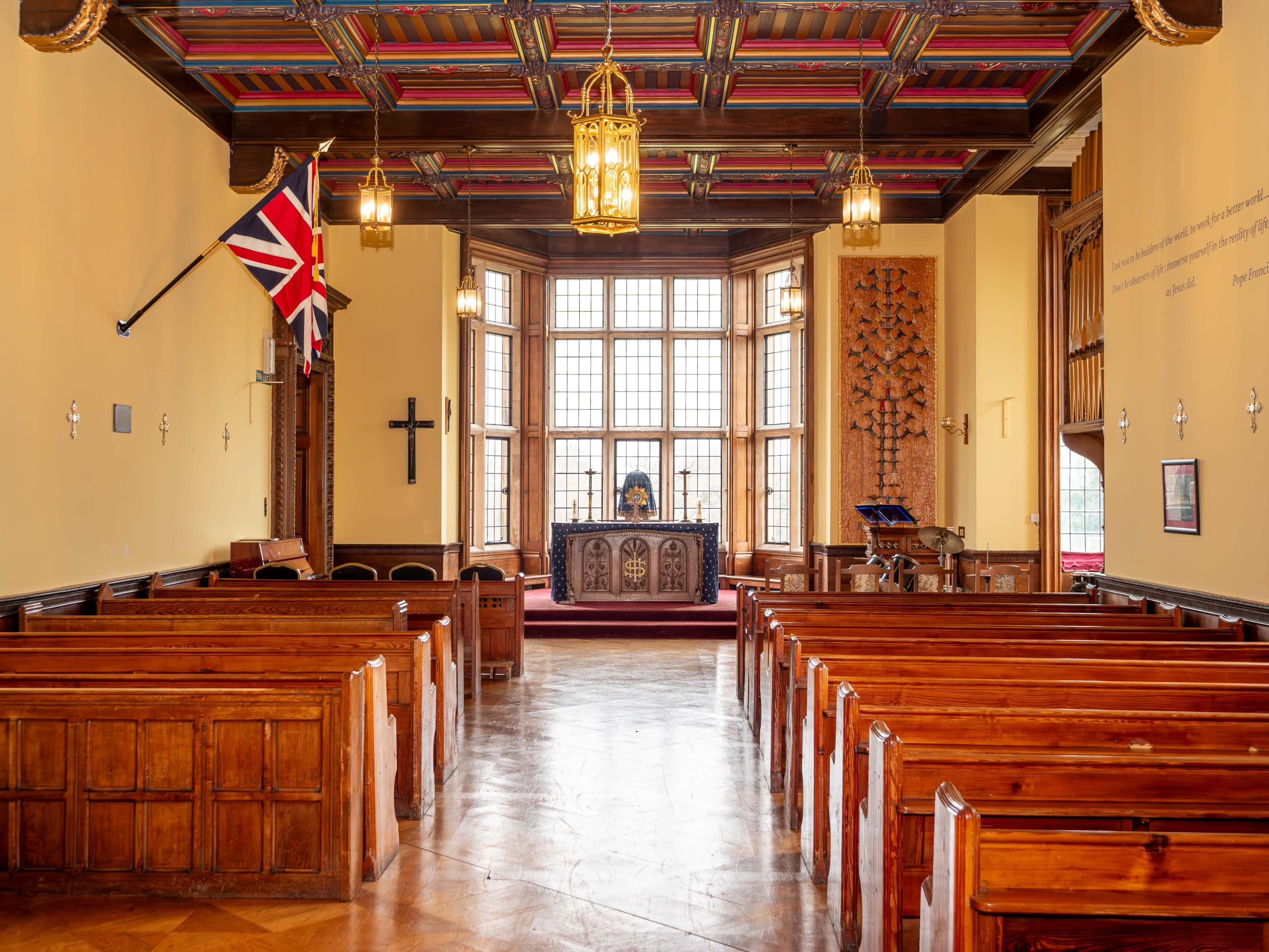The interior of a church features wooden pews facing a decorative altar beneath large windows, with a British flag displayed on the wall.