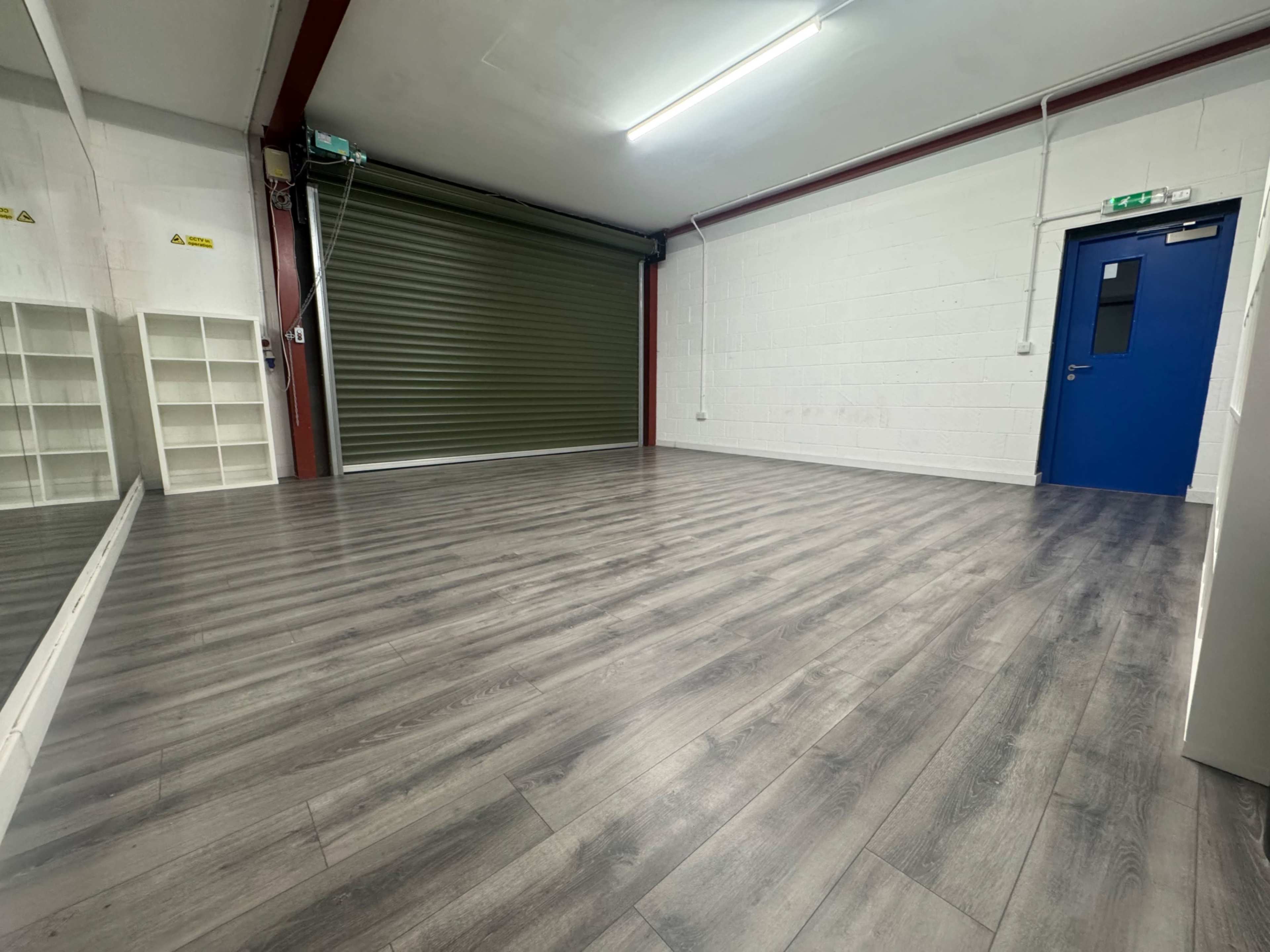 The image shows a spacious, empty room with wooden flooring, a green roll-up door, a blue door, and a white shelving unit against the wall.