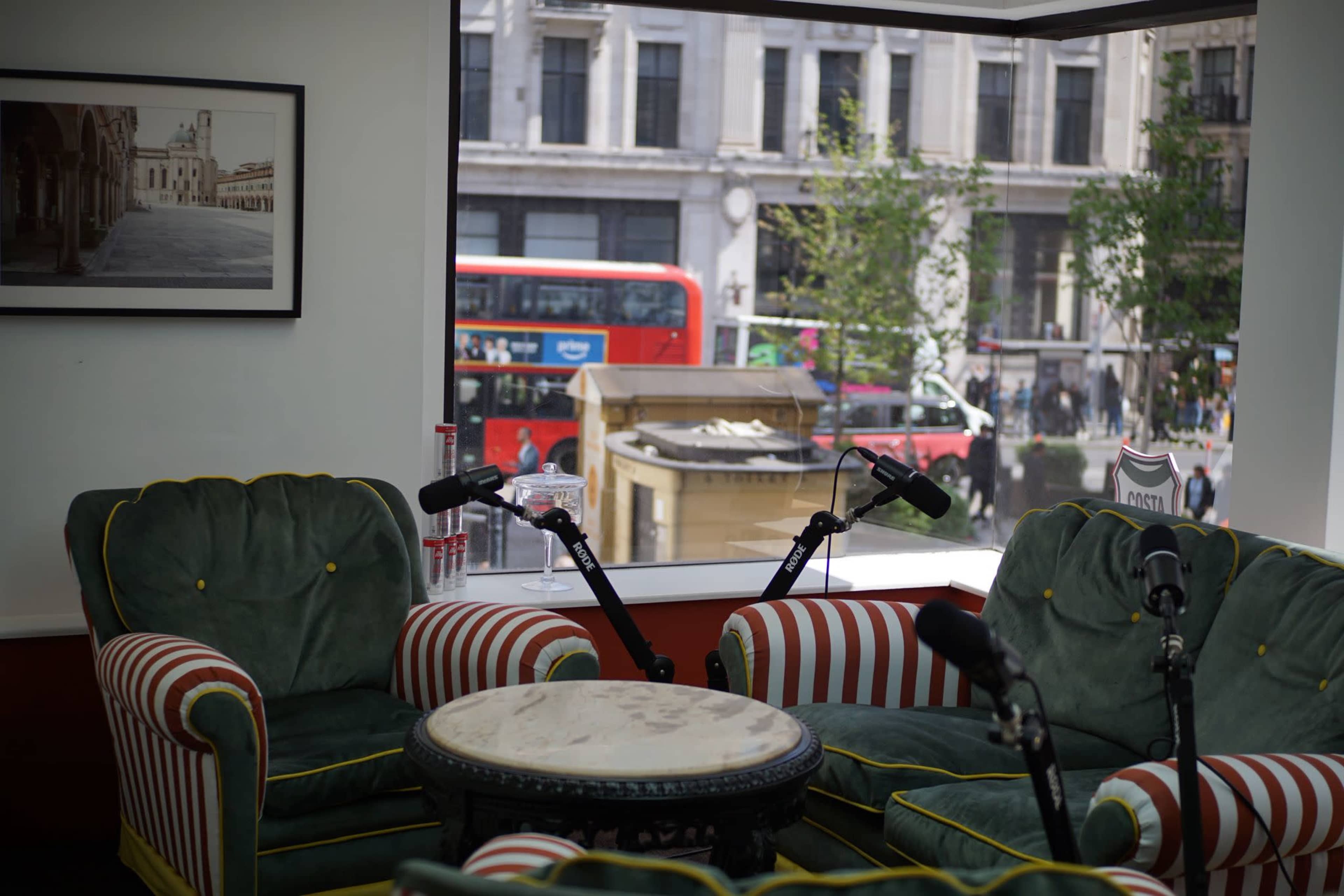 The image shows two green armchairs with striped red and white accents facing a small round table, positioned by a window overlooking a city street with a red double-decker bus passing by.