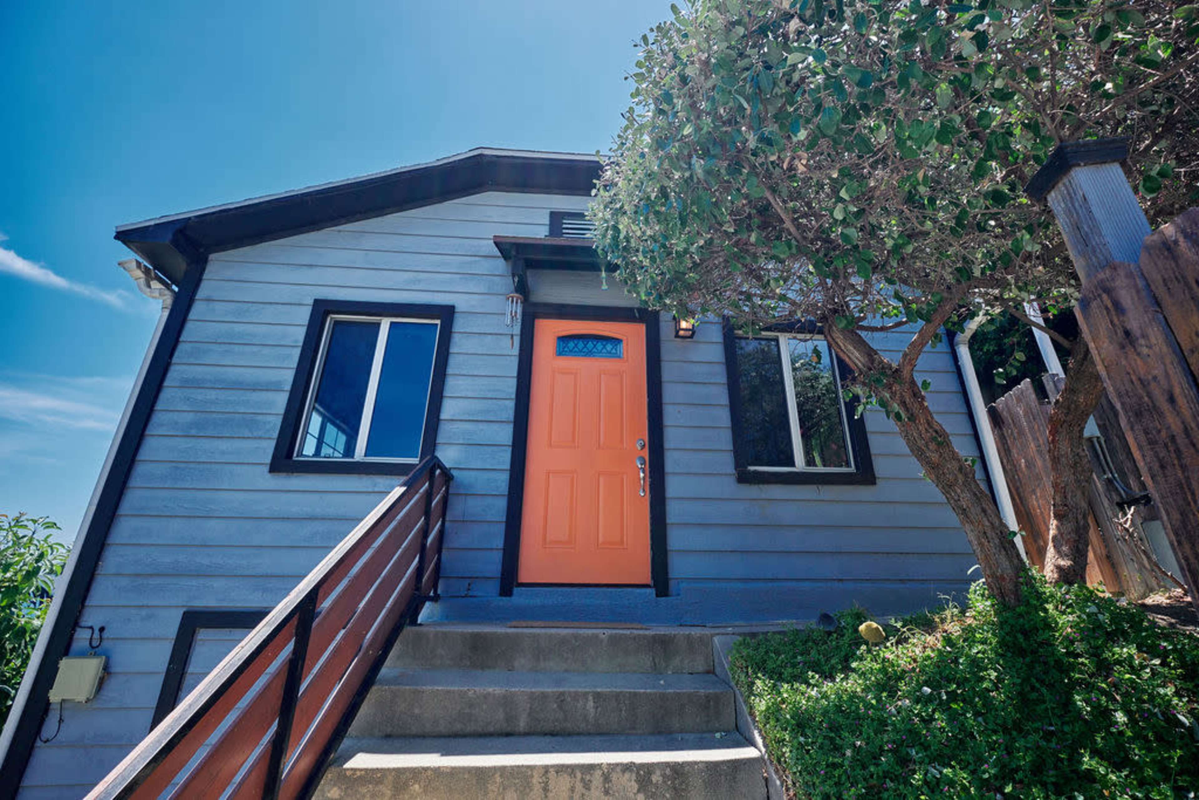 A light blue house features an orange front door and a sloped entrance with wooden steps.