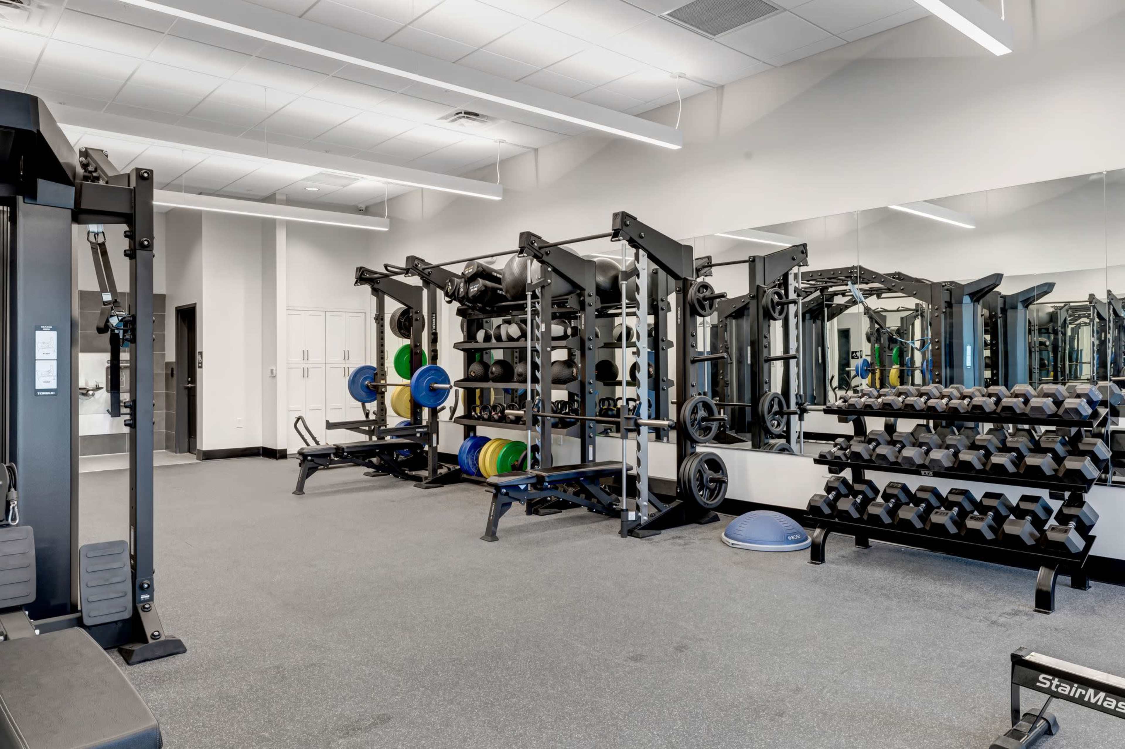 The image shows a well-equipped gym with weightlifting benches, dumbbells arranged on racks, and various exercise machines against mirrored walls.