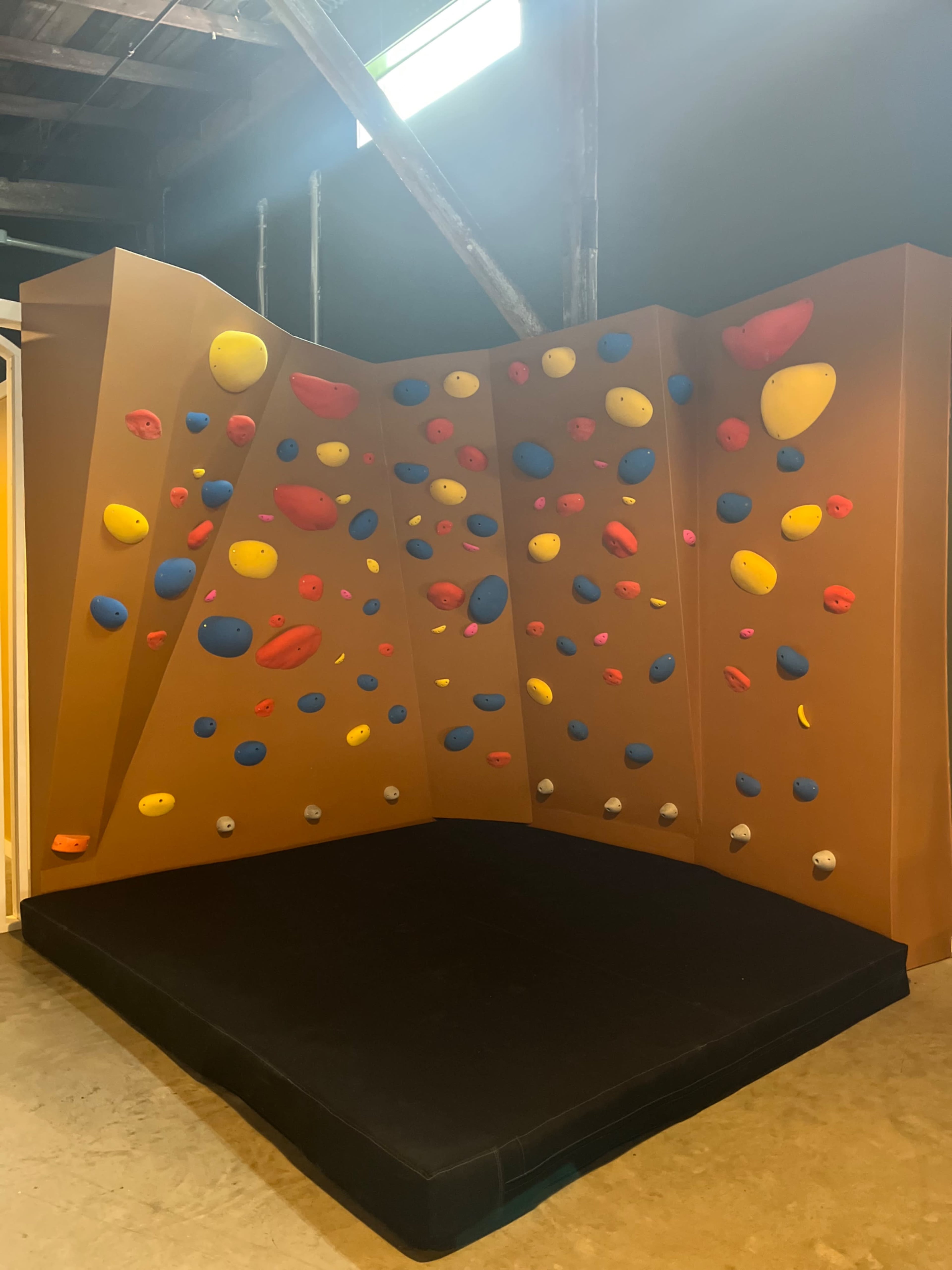 The image shows a bouldering wall with colorful holds in various shapes and sizes, positioned in a corner surrounded by a padded mat for safety.