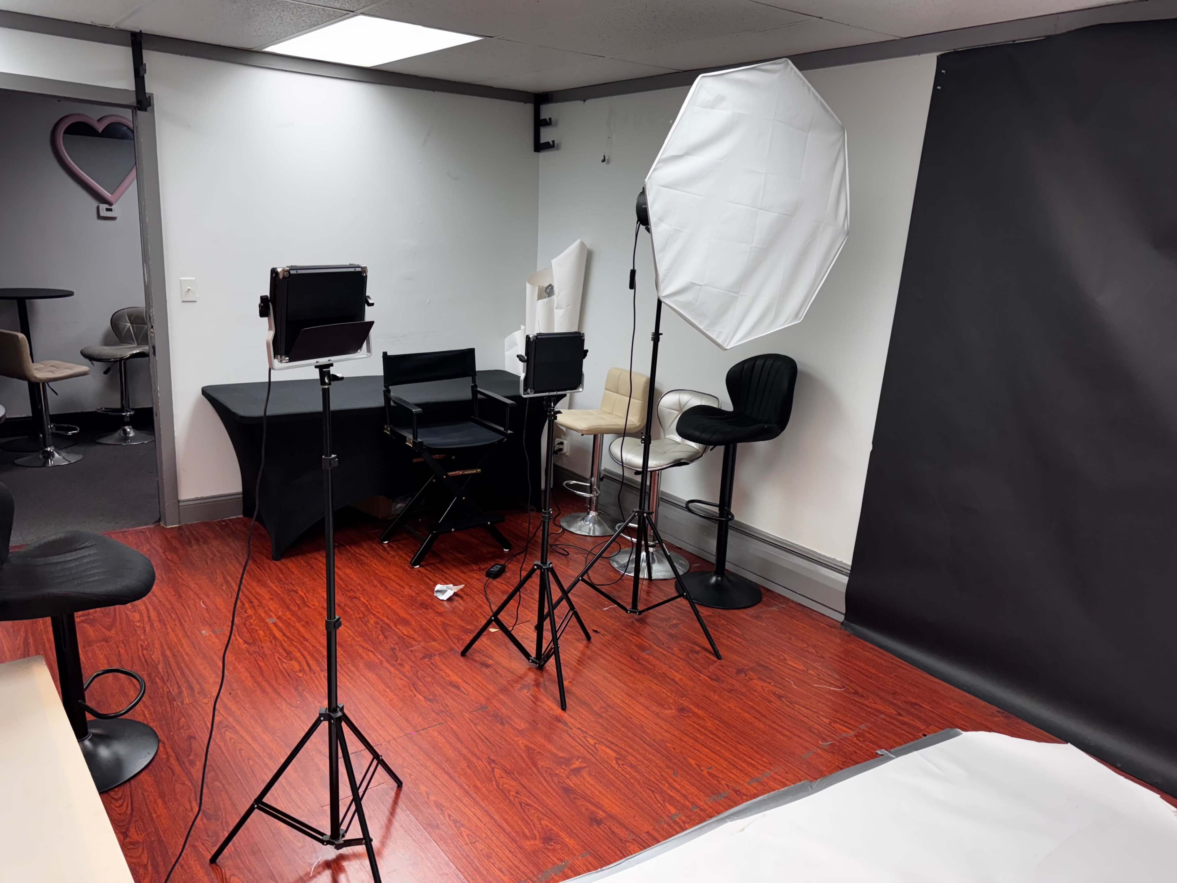 The image shows a photography studio setup featuring tripods with lights, a large softbox, a black backdrop, and various seating arrangements.