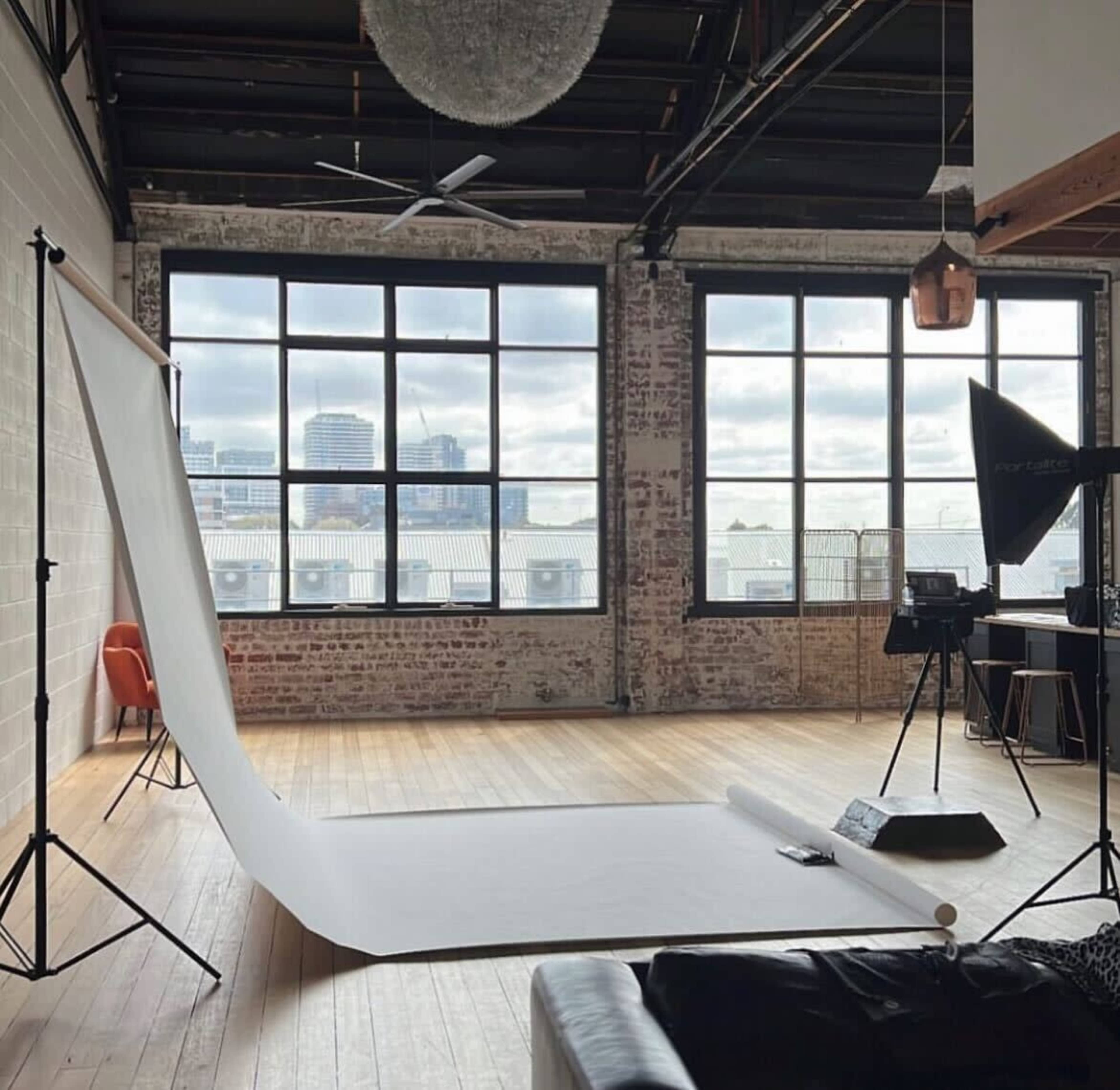 A photography studio with a white backdrop, large windows offering city views, and various lighting equipment set up beside a black couch.
