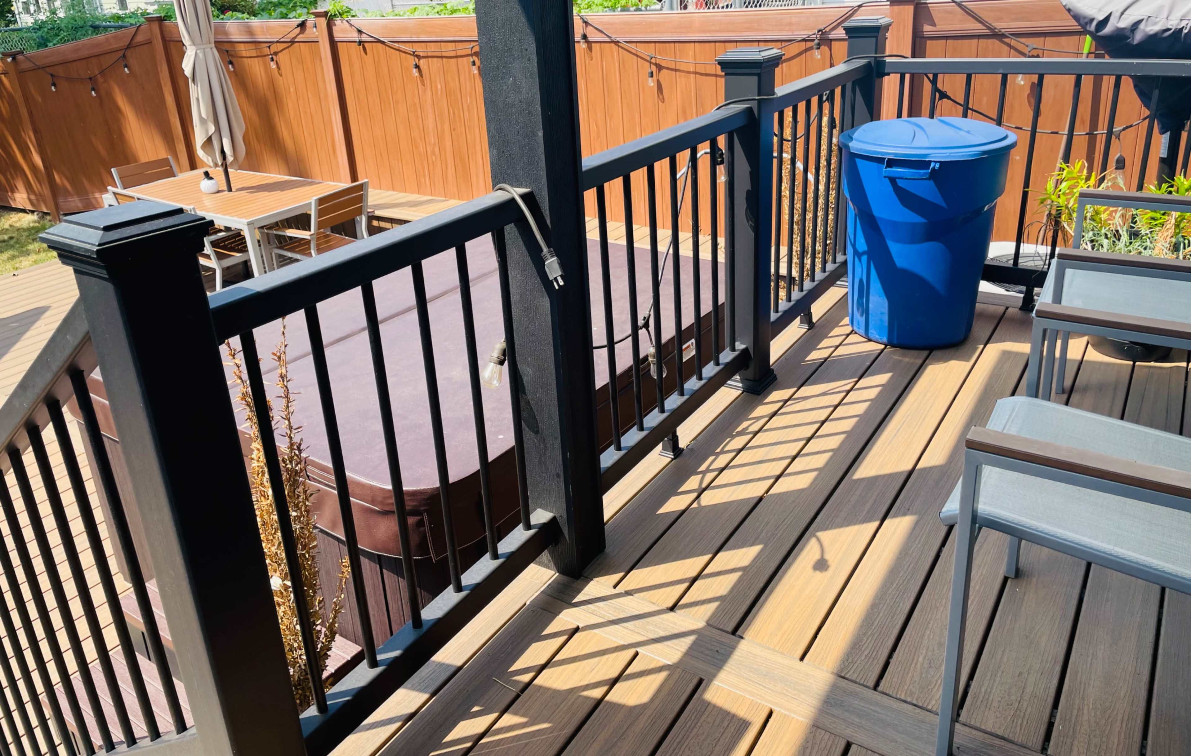 The image shows a deck area with a hot tub, a blue garbage bin, and outdoor furniture in a fenced backyard.