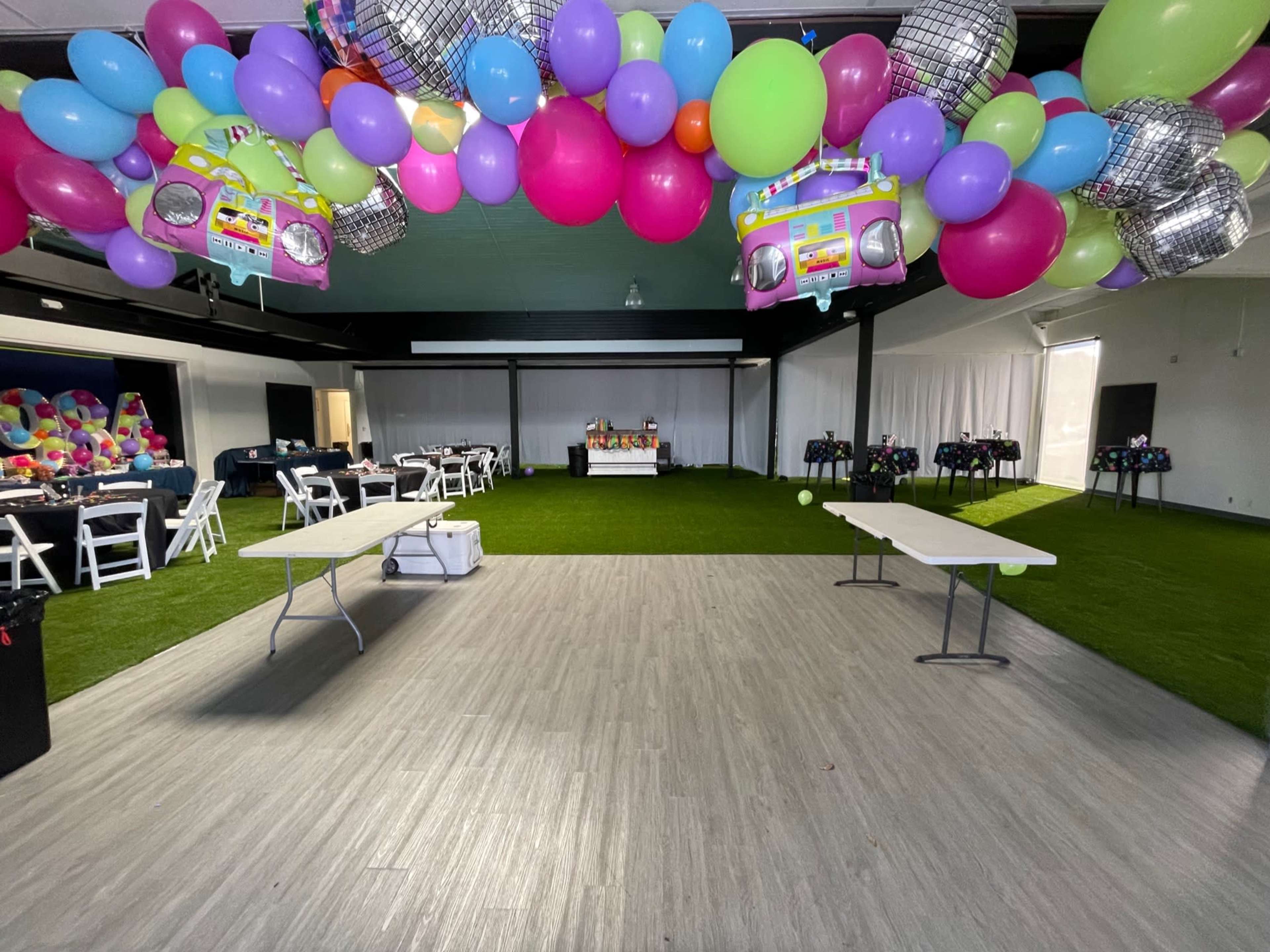 The venue features a decorated space with colorful balloons overhead, tables arranged for an event, and a grassy floor area.