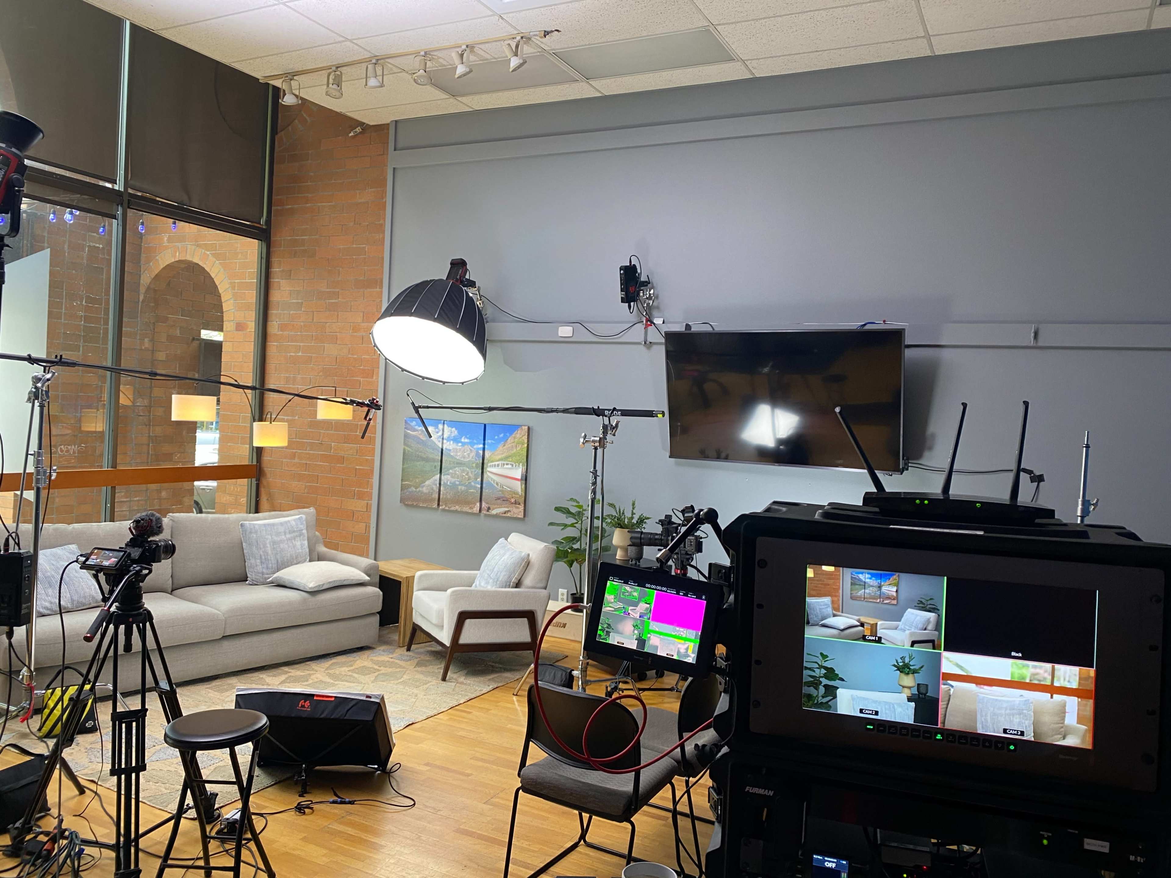 A video production setup in a studio with cameras, lighting equipment, and a seating area featuring a couch and a television screen.