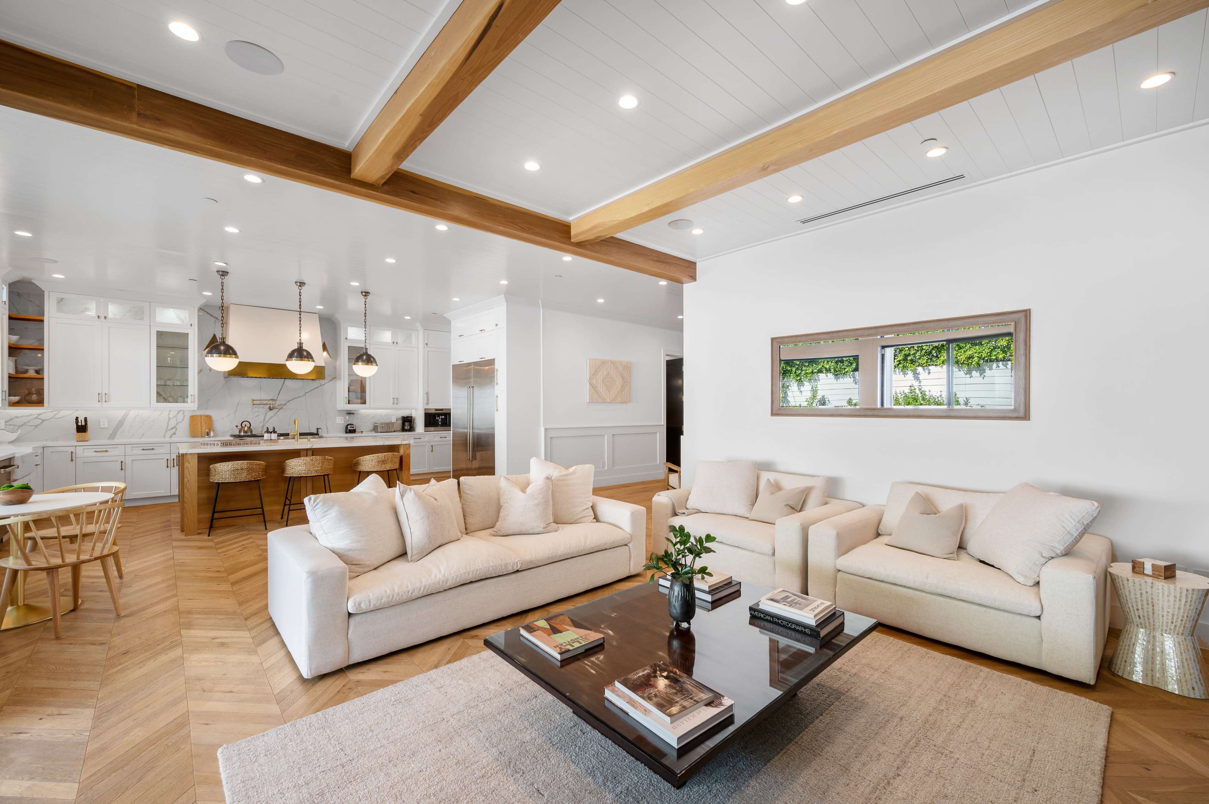 A bright, modern living room featuring two light-colored sofas facing a central coffee table, with a dining area and a white kitchen in the background.
