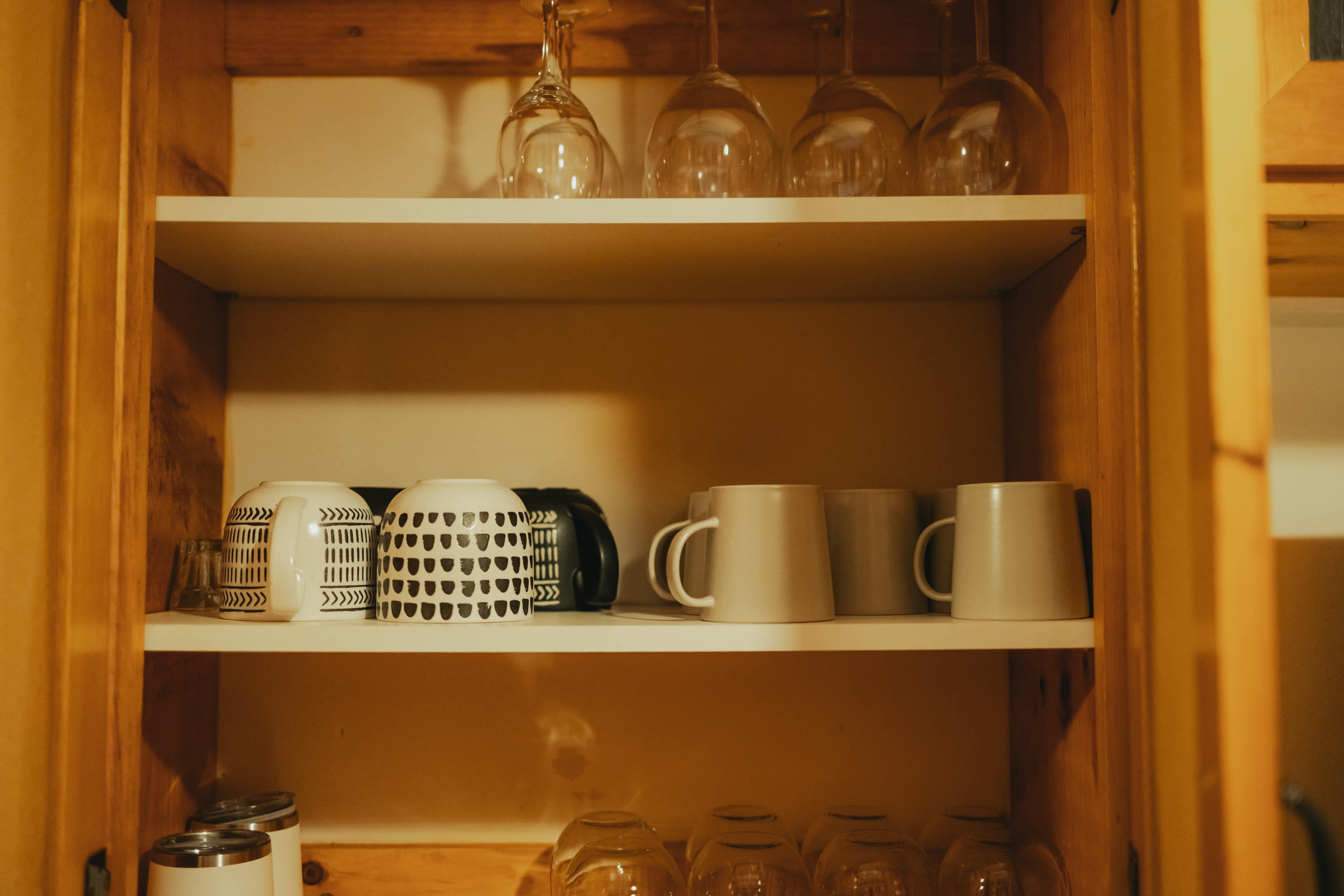 A wooden cabinet with two shelves containing a variety of mugs and glasses, including patterned and solid-colored designs.