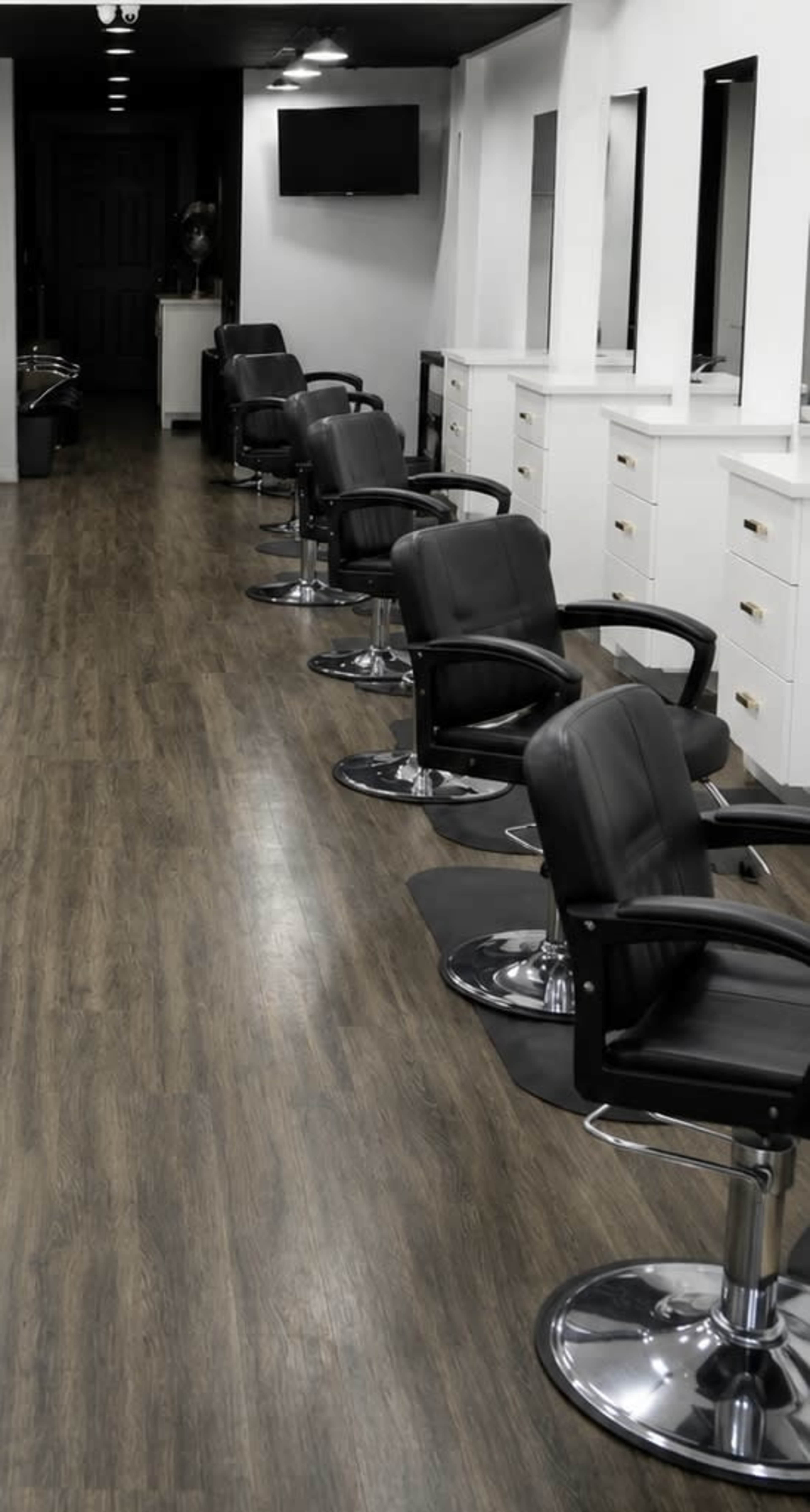 The image shows a row of five black salon chairs along a wooden floor in a well-lit hair salon.