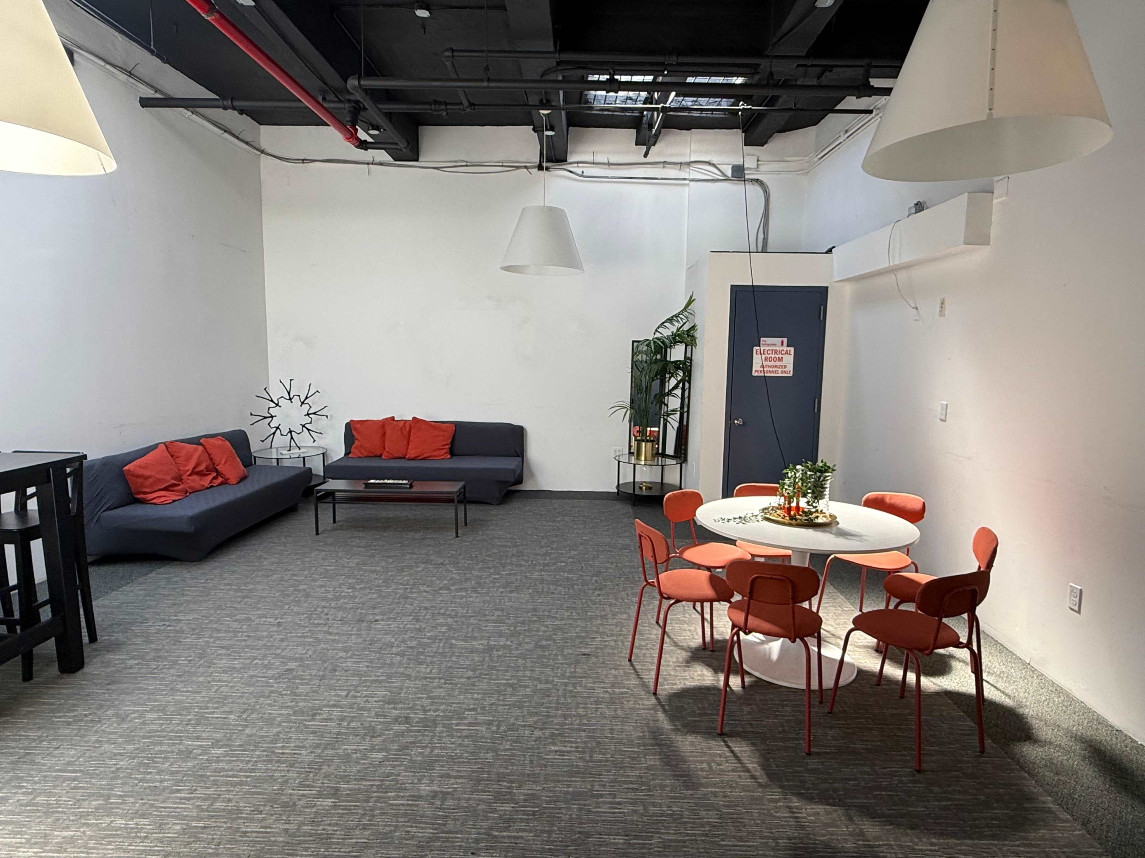 The image shows a spacious, minimally furnished room with a seating area featuring a dark sofa and red cushions, alongside a round dining table with eight red chairs.