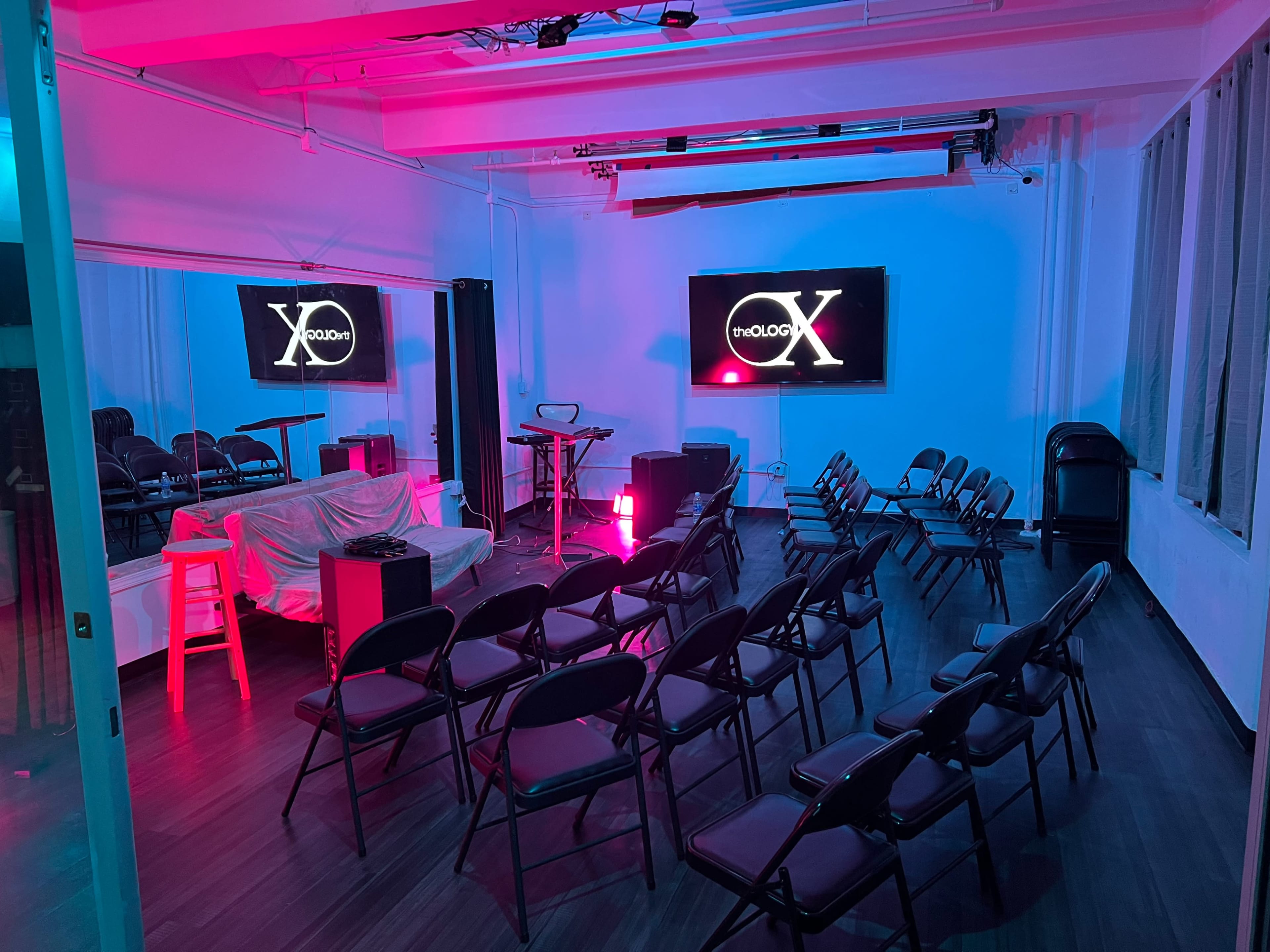 The image shows a dimly lit room set up with rows of chairs facing two screens and a small stage area.