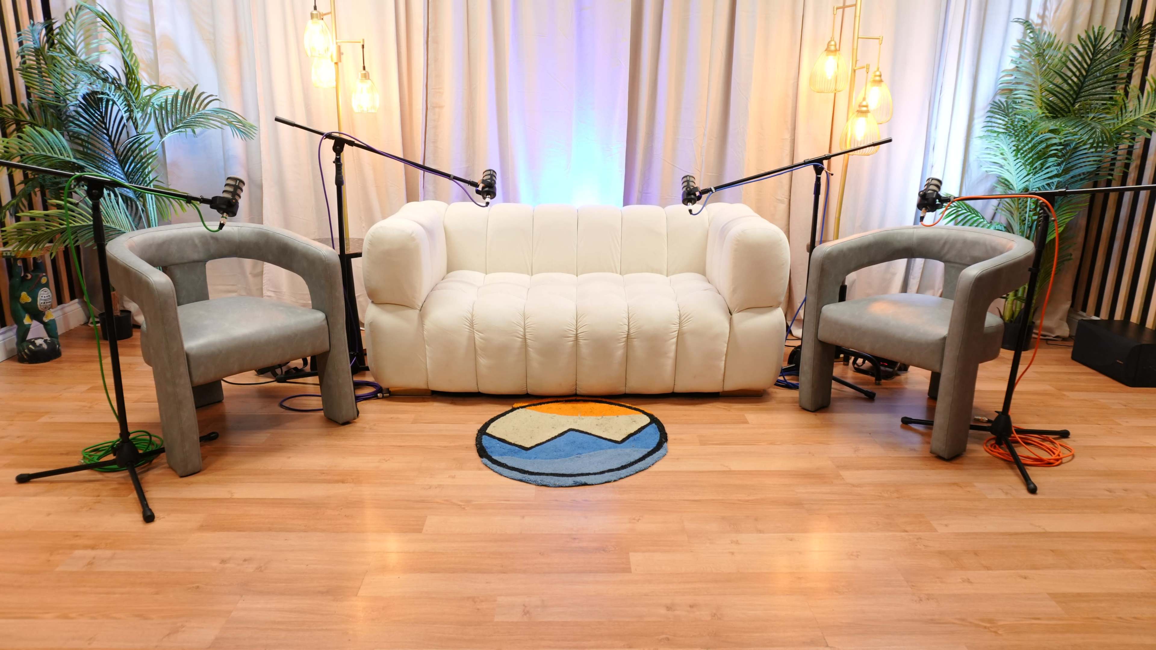 The image shows a minimalist podcasting setup featuring a white couch and two gray chairs, with microphones positioned nearby and a circular rug on the wooden floor.