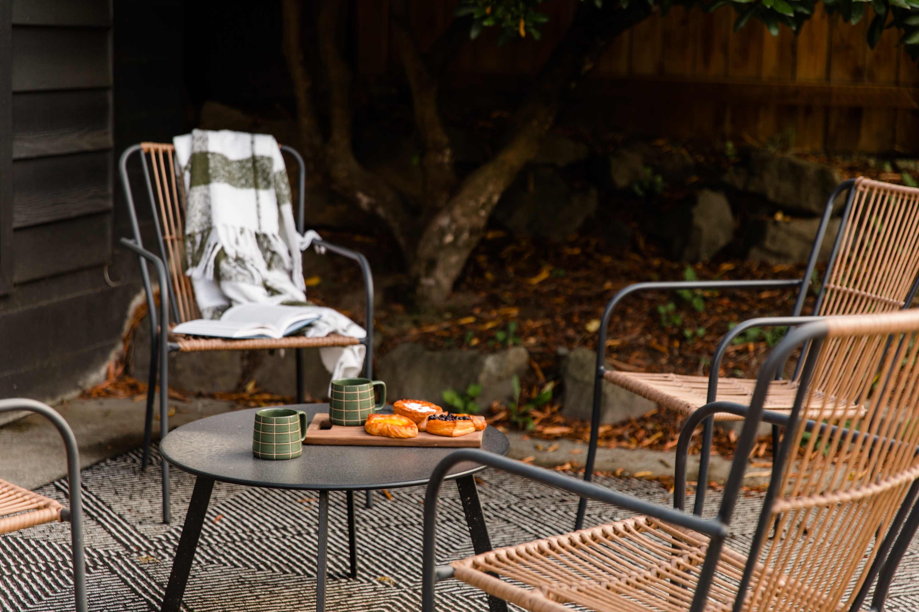 A cozy outdoor seating area features four chairs arranged around a small table with pastries and mugs, surrounded by natural elements.