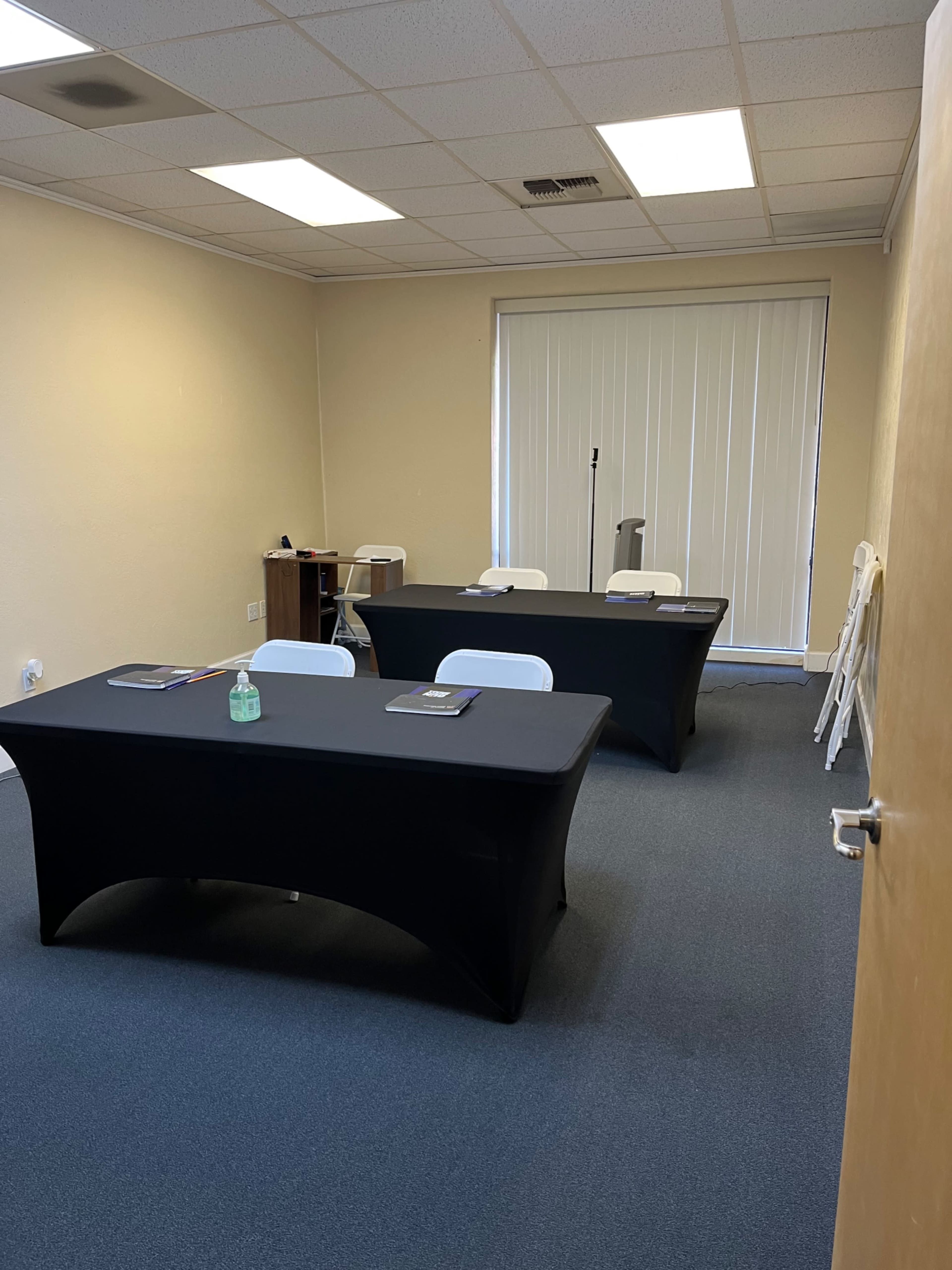 A small, empty meeting room with three black-covered tables, white chairs, and a window with vertical blinds.