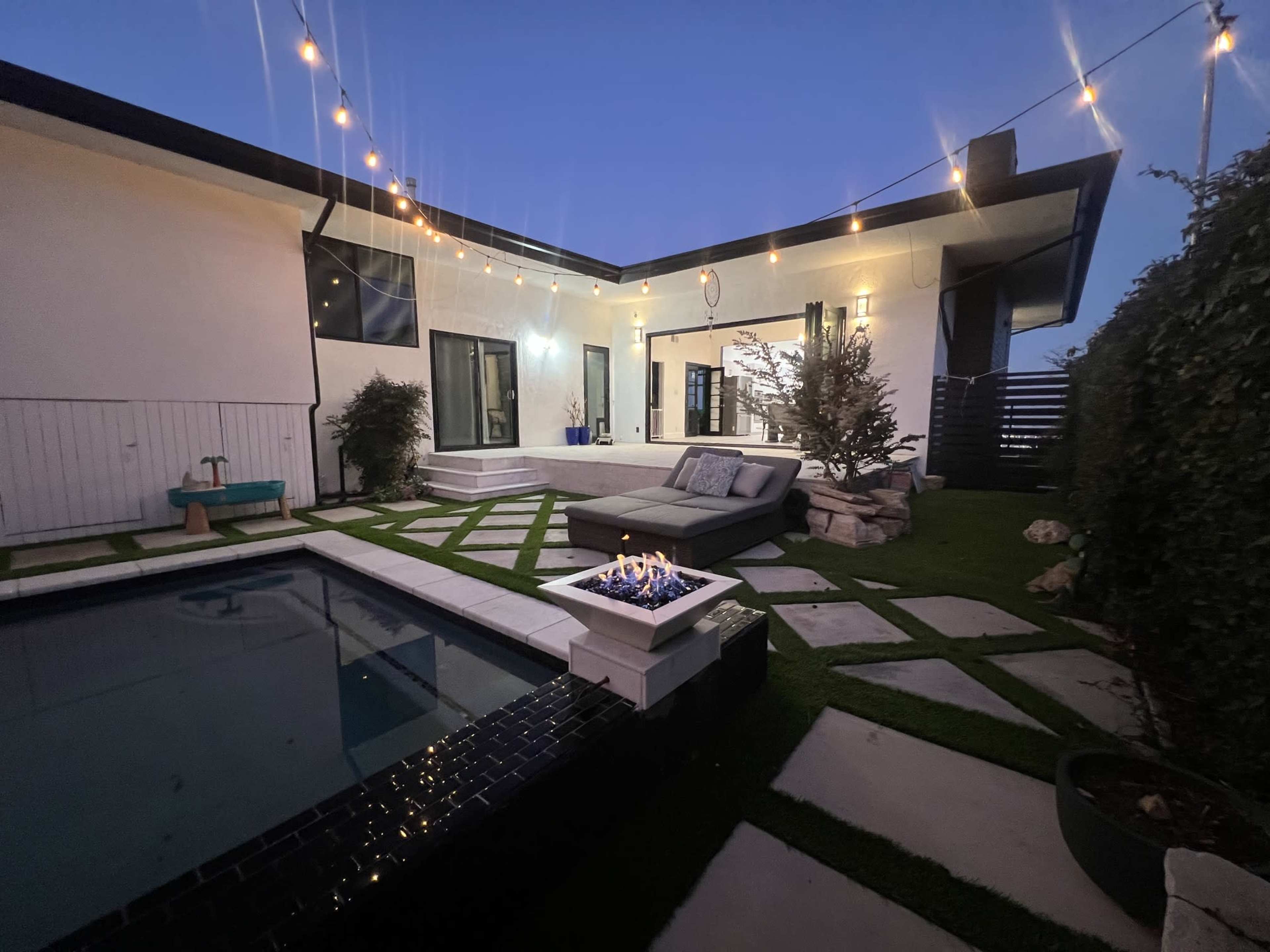 A modern backyard features a small pool, a fire pit with flames, a comfortable couch, and string lights above, creating an inviting outdoor space.